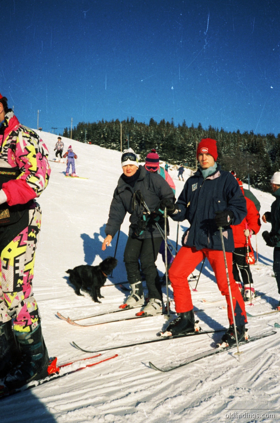 Vintage ski resort scene with three adults and a black dog on groomed slopes. Bright winter attire includes bold patterns (), red ski pants, and colorful ski jackets. Snow-covered pine forest and clear blue sky in background. Likely European alpine region.