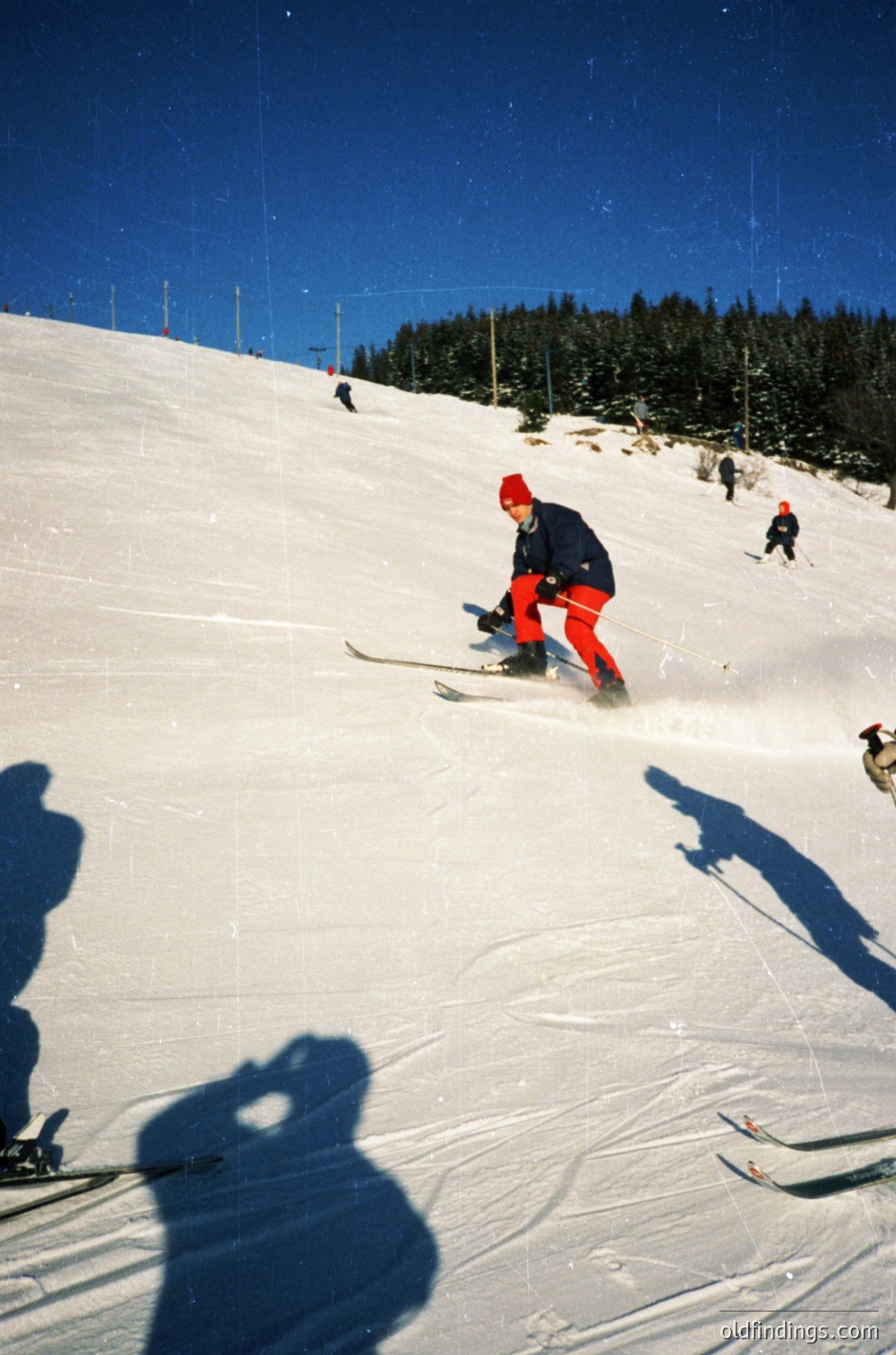 Vintage ski slope scene with skier in red cap and bright orange pants descending powdery snow. Other skiers visible in background on groomed trails. Clear skies and forested mountain backdrop suggest alpine resort. Likely mid-20th century winter sports photography.