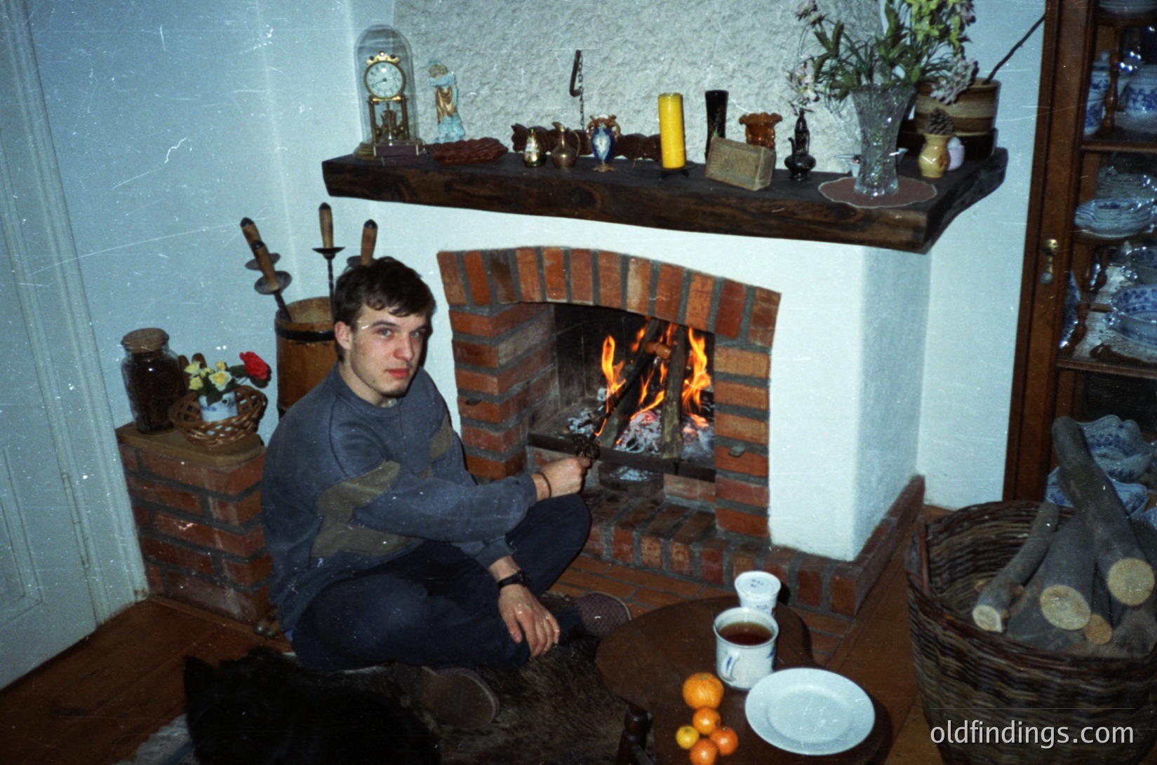 Vintage interior featuring a man seated by a brick fireplace with a warm fire. Surrounding decor includes brass candlesticks, ceramic jars, and a wooden shelf with vintage bottles. Coffee cup and saucer on a woven mat. Warm lighting and rustic wooden floor enhance cozy ambiance.
