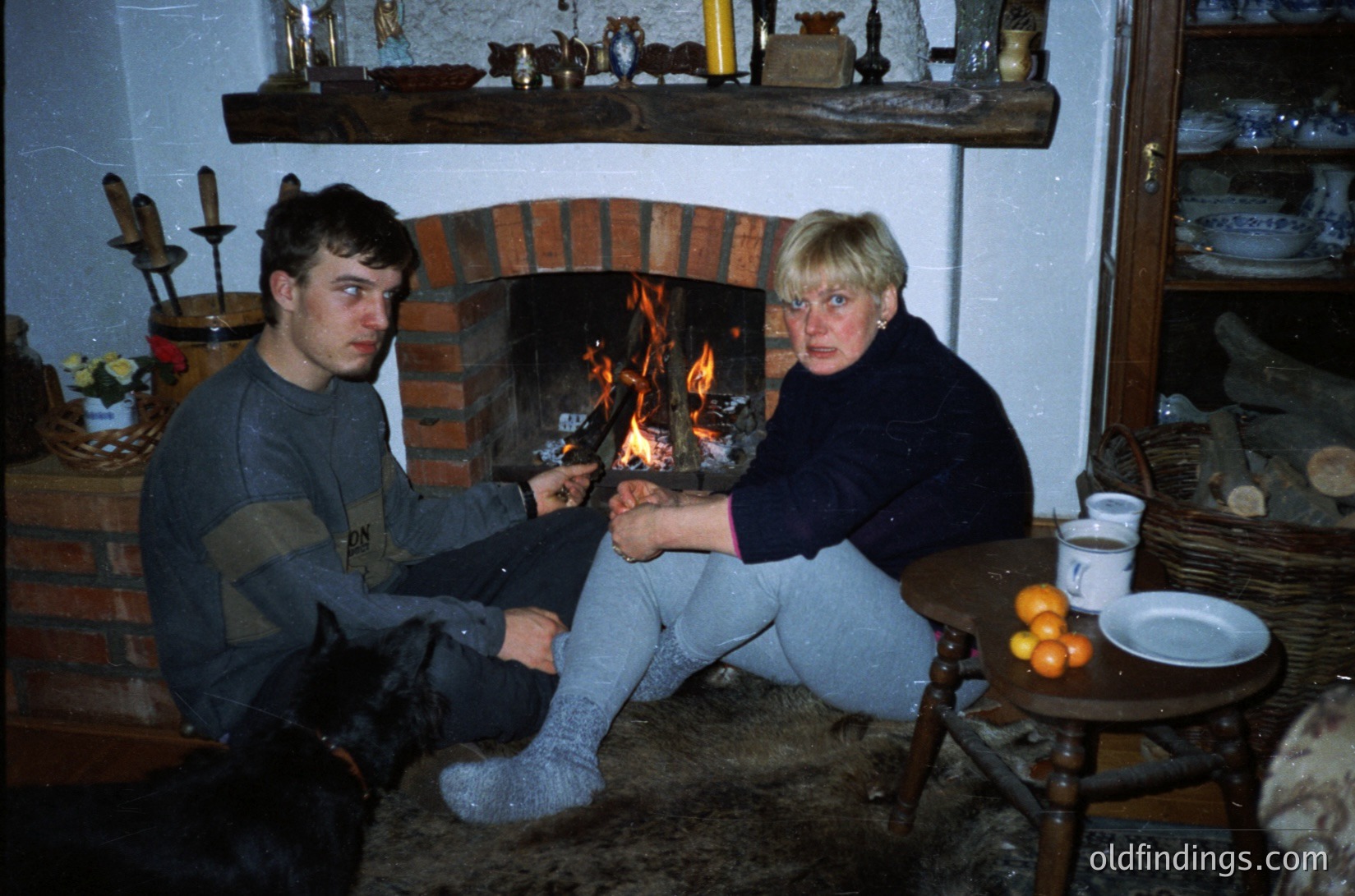 Two individuals seated by a brick fireplace in a rustic interior, likely Eastern European . The man wears a striped sweater and light pants, while the woman in a dark sweater and light trousers holds a small object near the fire. Surrounding decor includes ceramic figurines, woven baskets, and a small table with a bowl of fruit. Warm, intimate domestic scene.