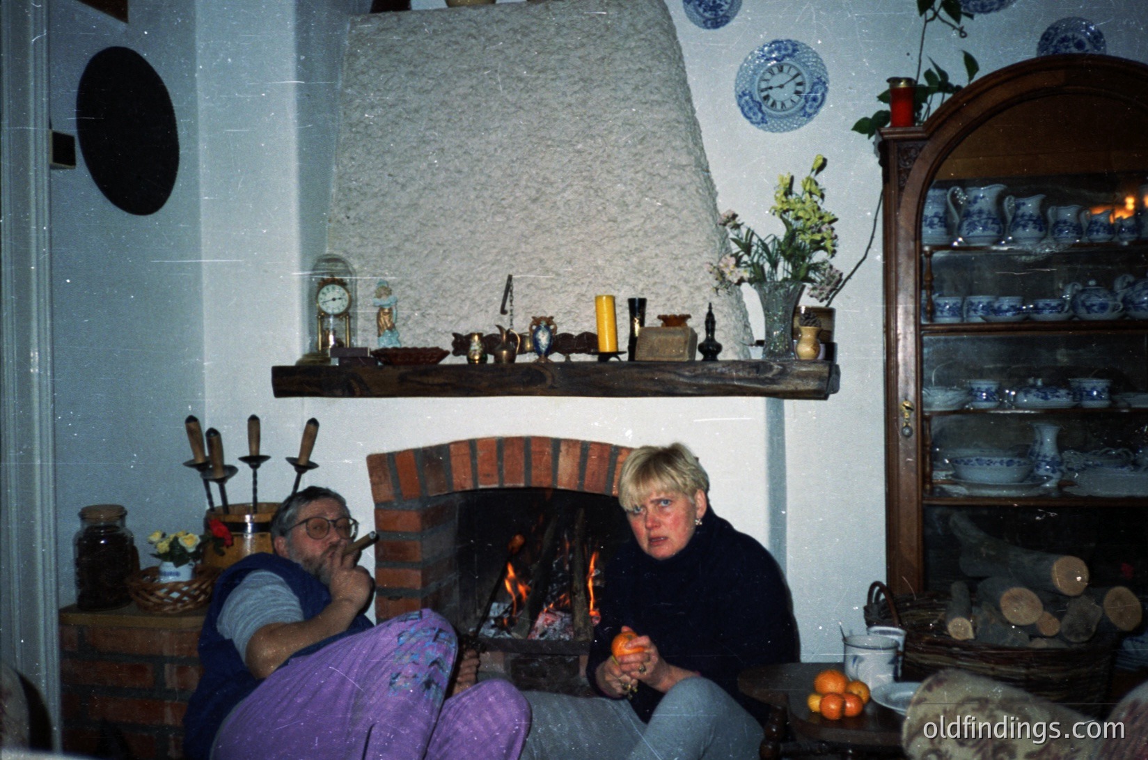 Two adults seated by a brick fireplace in a cozy, rustic interior. Wooden shelves display ceramic jars, blue-and-white patterned dishes, and a vintage clock. Warm light from the fire illuminates the scene, suggesting a 1970s-1980s European countryside home.