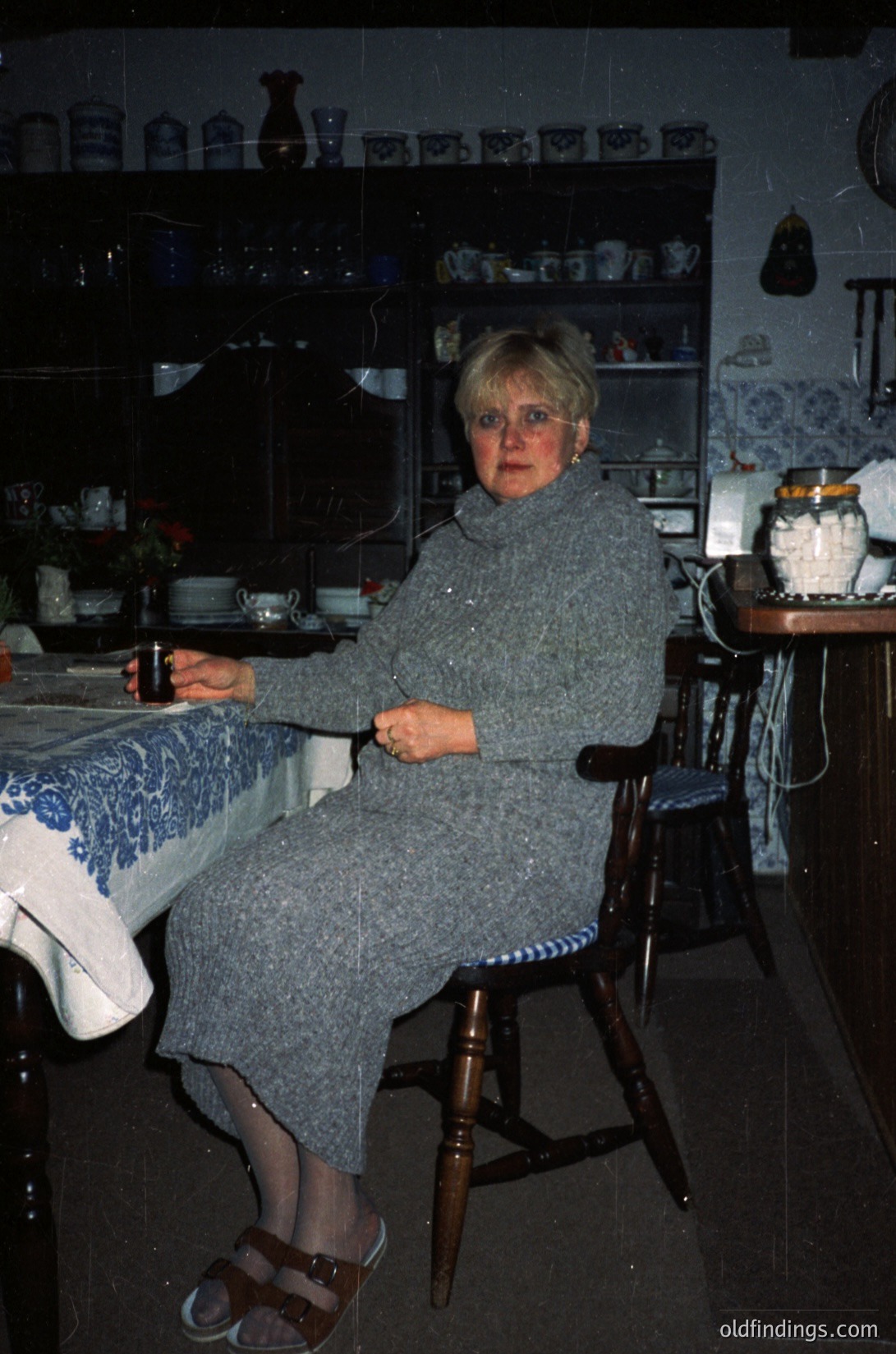 Vintage kitchen interior featuring a woman in a 1970s-style gray knit dress and sandals, seated at a wooden table draped with a floral-patterned tablecloth. Shelves display ceramic jars, plates, and decorative items. Warm lighting and retro decor evoke mid-century domestic life.