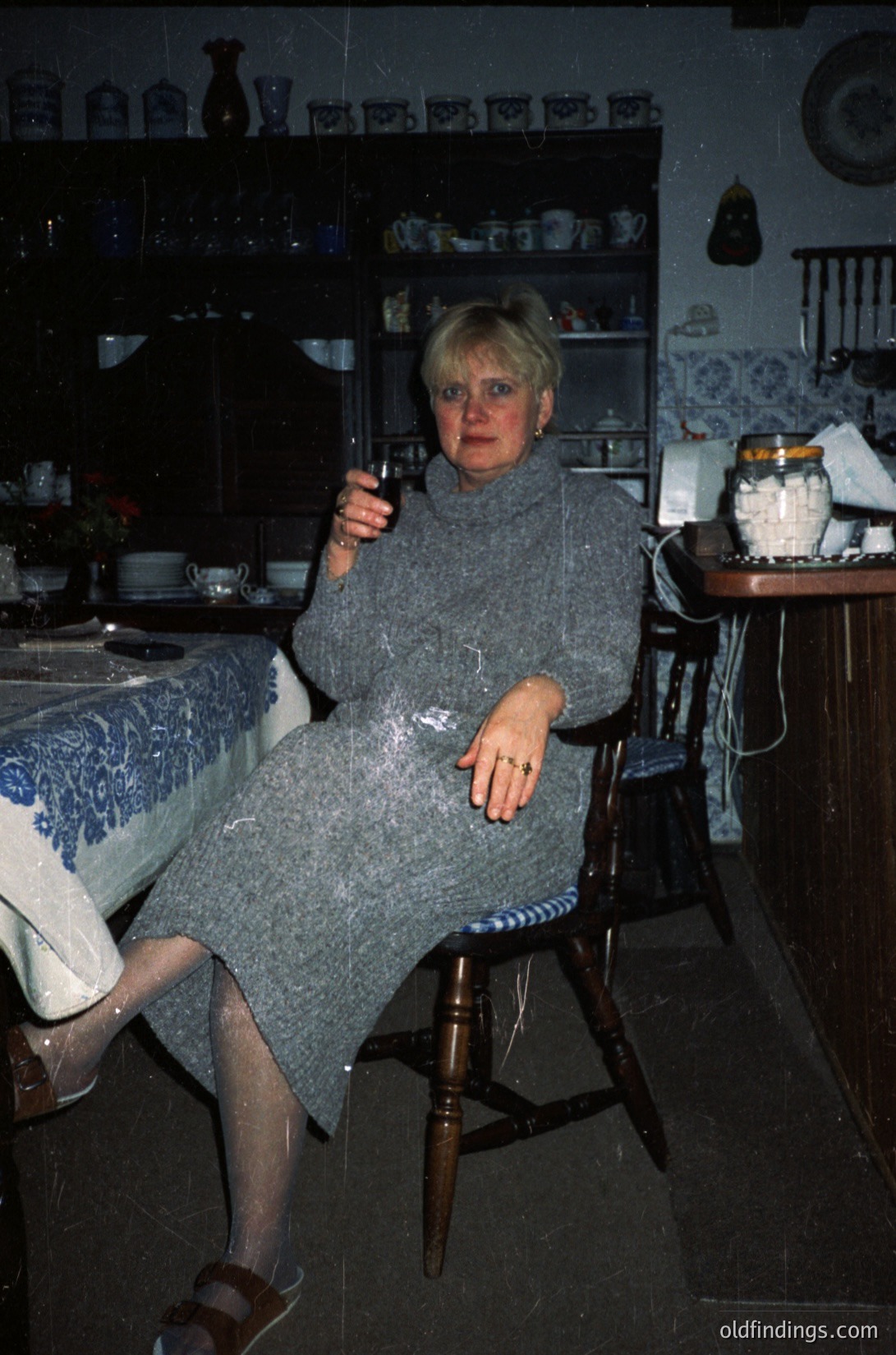 Mid-century kitchen interior featuring a woman in a textured gray dress, seated on a wooden chair. She holds a wineglass, surrounded by vintage ceramic dishes and patterned tableware. Warm lighting and a cozy, lived-in atmosphere suggest a 1970s European home. écor