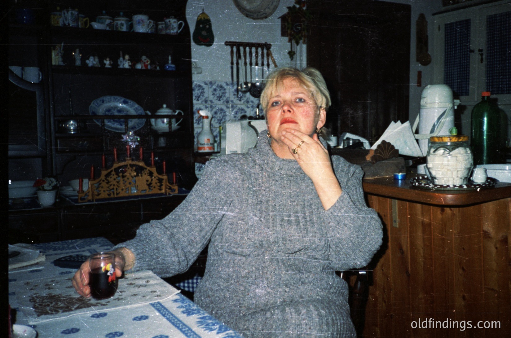 Vintage kitchen scene featuring a woman in a textured sweater, seated at a table adorned with a blue-and-white tablecloth. She holds a lit candle in a dark holder, while a teapot and decorative plates rest on shelves behind her. Warm, ambient lighting highlights ceramic figurines and a hanging lamp. Likely Eastern European domestic interior, 1980s–1990s.