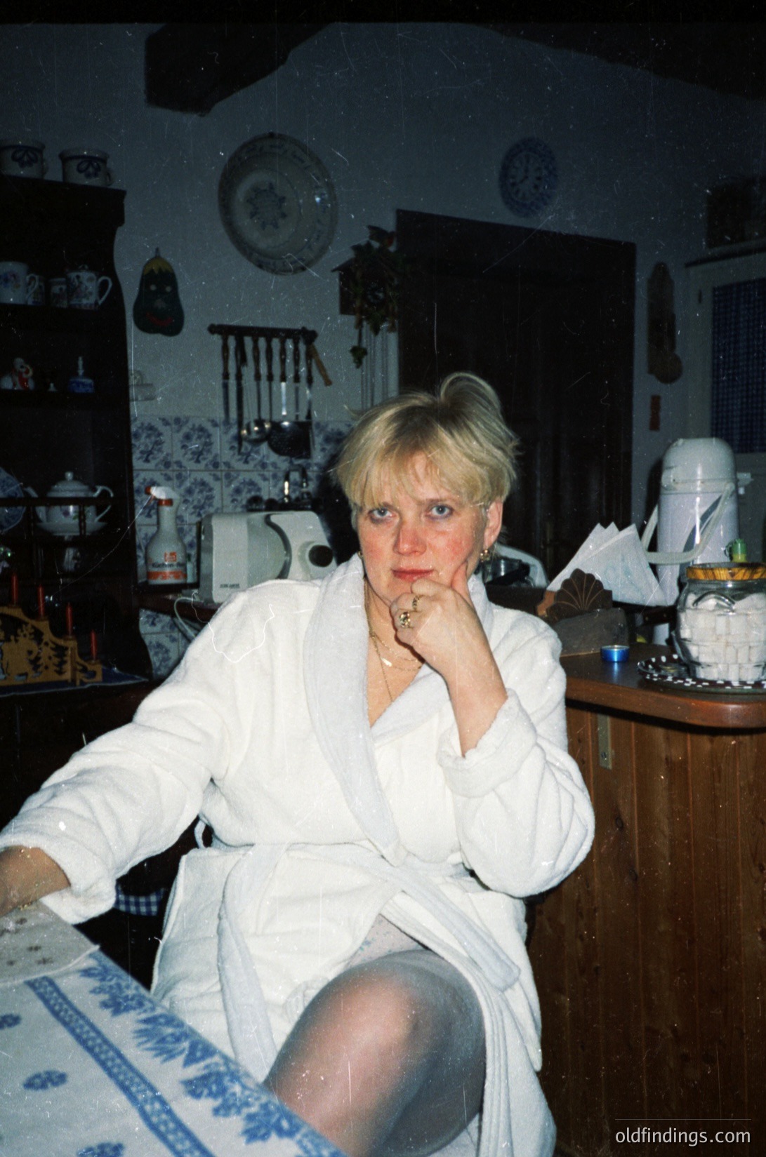 Vintage kitchen interior featuring a woman in a white robe, seated at a wooden table with blue-and-white patterned cloth. Surrounding her: ceramic teapots, vintage kitchenware, and a retro radio. Likely late 20th century (1970s–1990s).