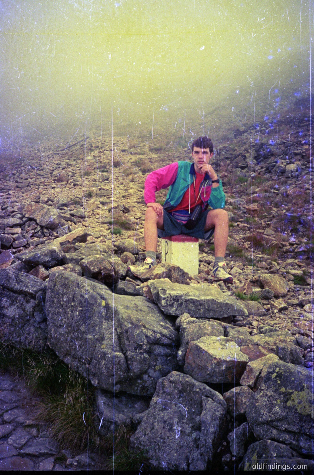 Hiker in vintage outdoor gear poses on rocky alpine trail, misty backdrop suggests high elevation. Brightly colored jacket and shorts hint at 1970s–1980s hiking fashion.