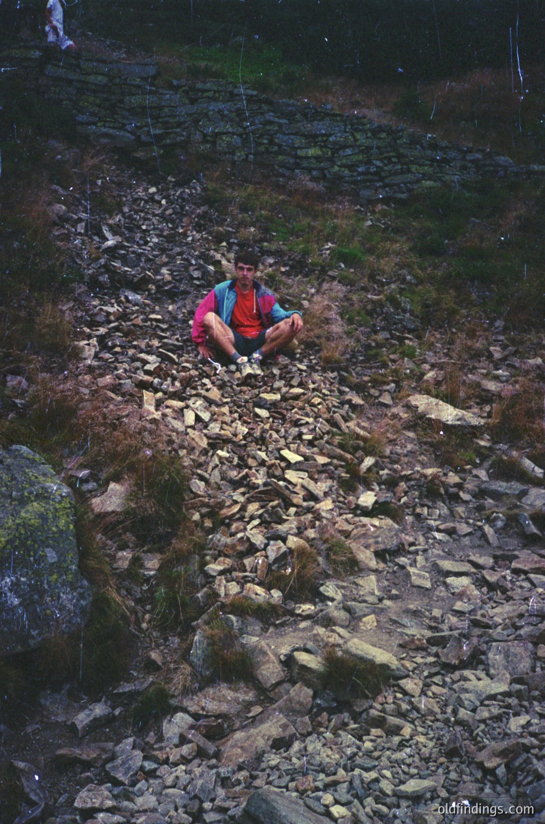Individual sits on rocky alpine trail, wearing bright red jacket and blue backpack. Stone steps and mossy terrain suggest a rugged hiking path. Likely mid-20th century outdoor photography.