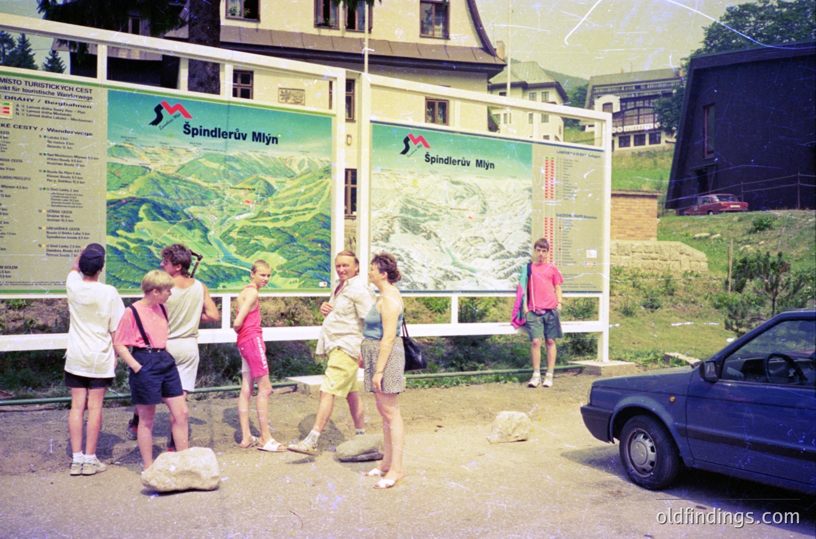 Vintage 1970s alpine tourist scene at **Spindlerův Mýtň** (Czech Republic). Five hikers in retro outdoor wear examine detailed trail maps beside a parked car. Green signage highlights mountainous terrain with ski routes. Wooden chalet-style buildings and lush greenery frame the scene.