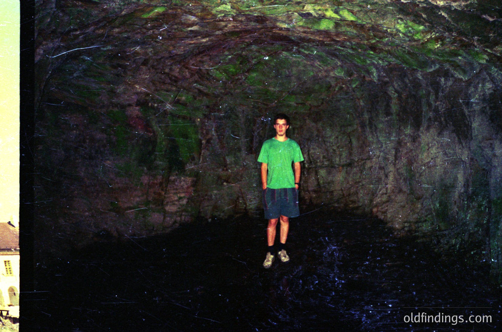 A young individual stands in a narrow, moss-covered rock crevice with water at the base, likely a natural cave or grotto. The person wears a green T-shirt, dark shorts, and sandals. The lighting suggests an overcast or shaded environment, enhancing the rock’s textured, organic appearance.