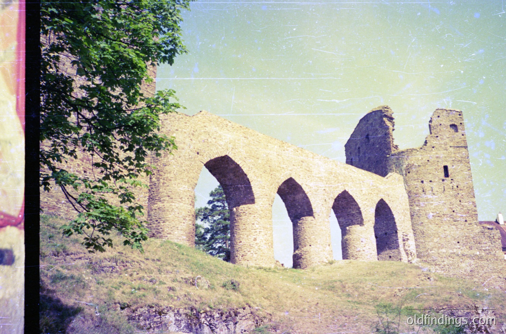 Vintage photograph of a medieval stone ruin featuring three arched doorways and a tower remnant, likely Byzantine or early Christian architecture. Overgrown greenery frames the structure, suggesting rural abandonment. Film grain and sepia tone indicate 20th-century analog capture.
