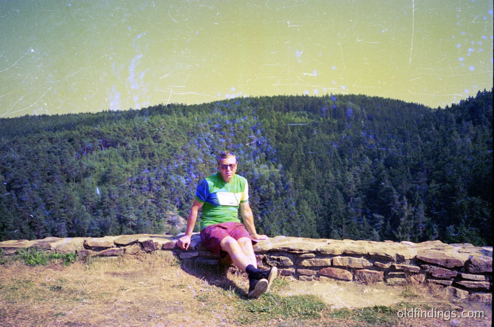 Vintage photo of a man seated on a stone wall overlooking dense forested hills, likely Eastern Europe. His striped shirt and shorts suggest mid-20th century fashion. The faded, yellow-tinted film enhances nostalgic appeal.