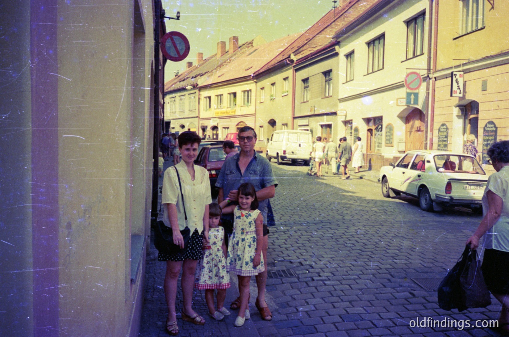A family of four poses on a cobblestone street in a European town, likely 1960s–1970s. The woman wears a short-sleeve blouse and skirt; the man, a button-down shirt and cap. Two children in floral dresses stand beside them. Vintage cars and classic street signs () frame the scene. Warm, vintage filter enhances nostalgic charm.