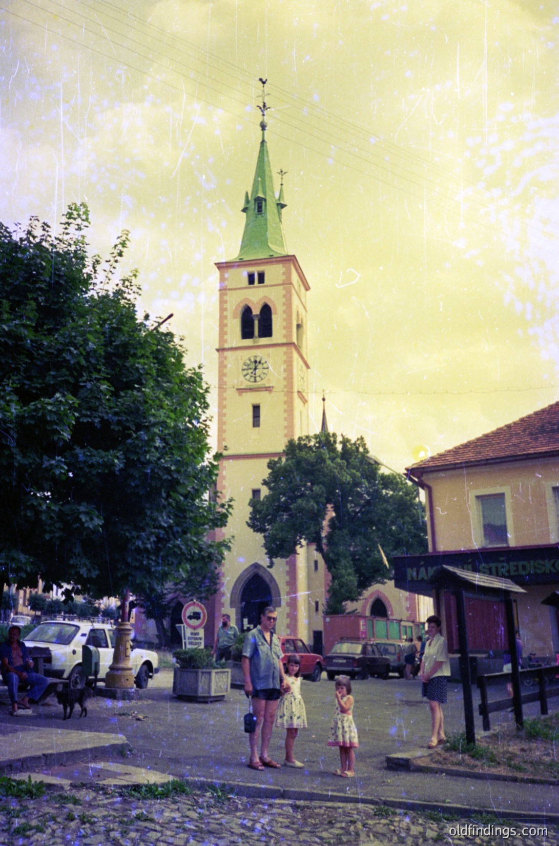 Vintage street scene featuring a prominent **19th-century church tower** with a green spire and clock, framed by cobblestone streets and lush trees. Three adults and a child stand near a small fountain, dressed in mid-20th-century attire. Vintage cars and a parked truck add to the nostalgic atmosphere.