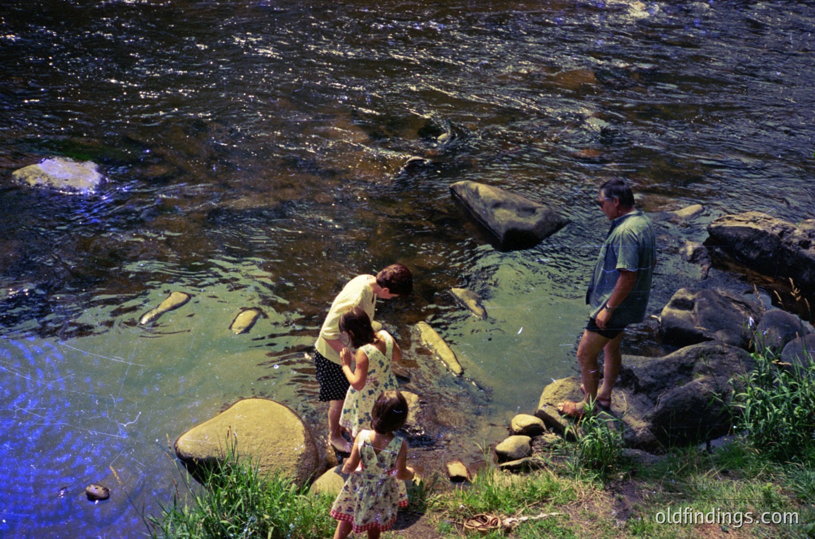 Family of three wading in shallow, rocky river—adults assist child in water. Mid-20th century clothing suggests or . Natural setting with clear water and greenery. Ideal for outdoor lifestyle, travel, or historical research.