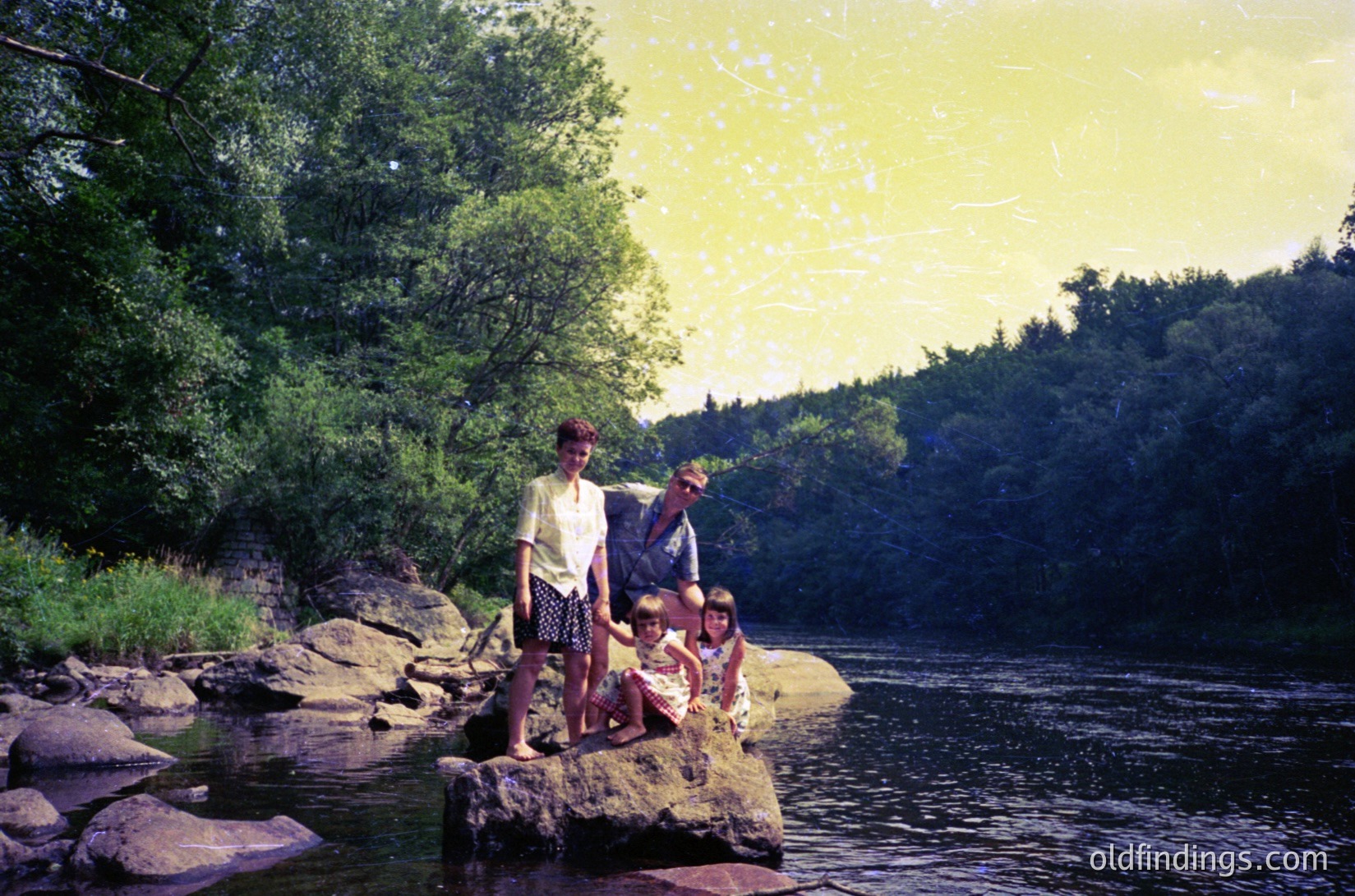 Three individuals pose on river rocks during golden-hour lighting, mid-1970s. Woman in patterned skirt and blouse stands beside two children in casual summer attire. Lush greenery and dense forest frame the serene riverbank. Vintage sepia-toned photo captures mid-century outdoor family life.