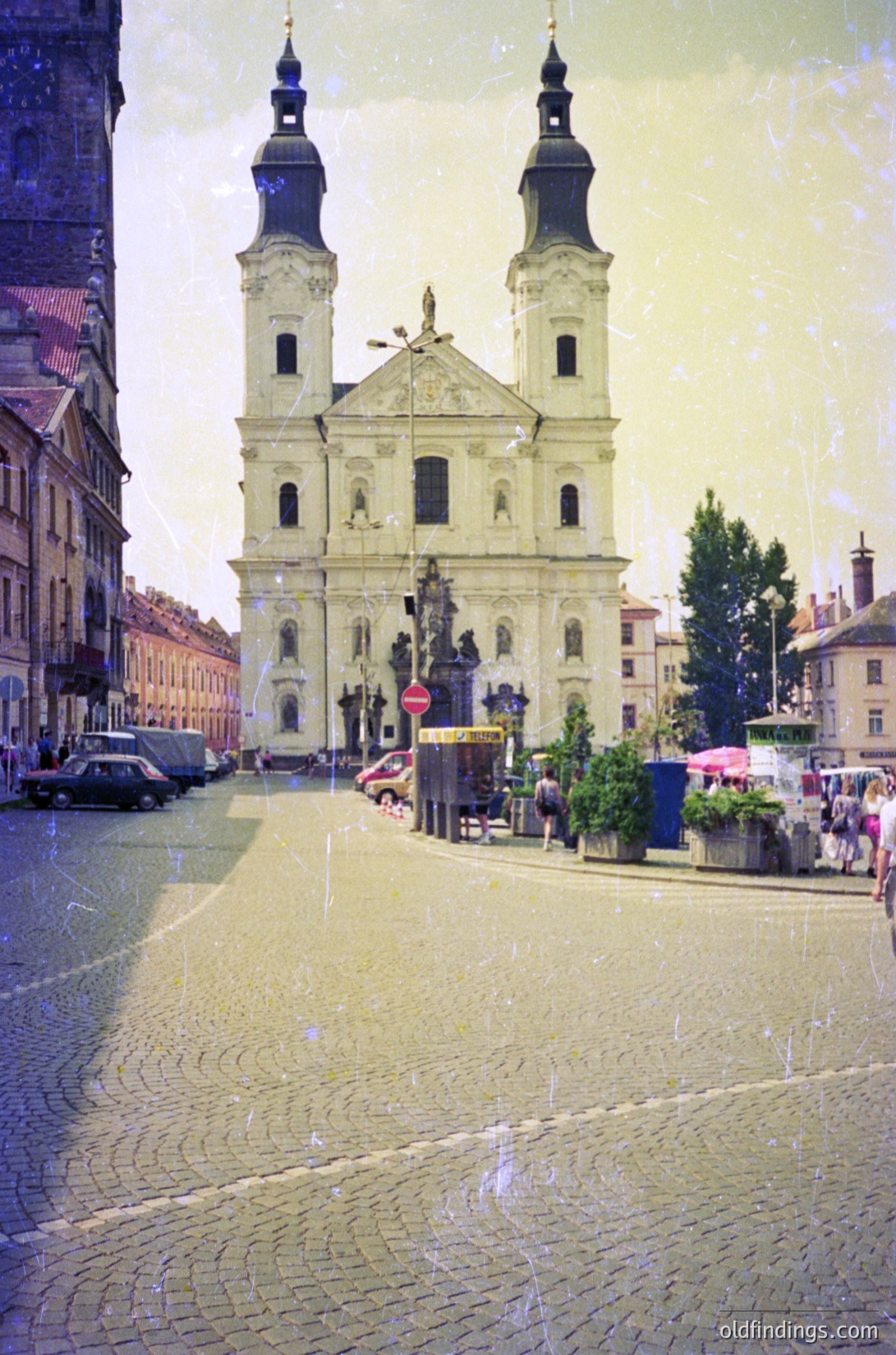 Baroque-style church with twin towers and ornate façade, likely 18th-century European urban design. Cobblestone plaza with wet surface, indicating recent rain. Surrounding buildings feature classic European architecture. Pedestrians and parked cars suggest active city life.