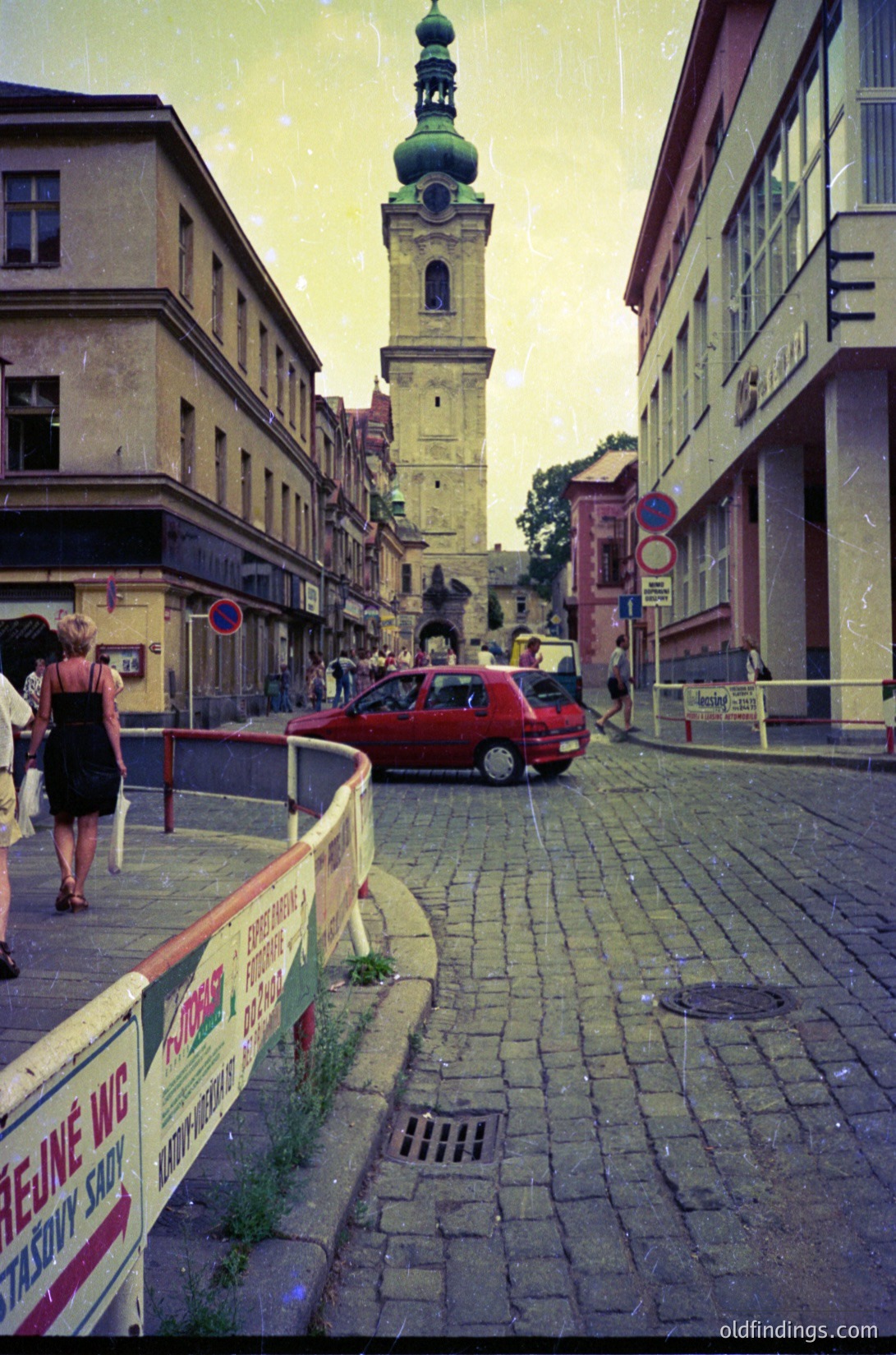 Vintage street scene featuring a cobblestone road leading to a prominent **18th-century bell tower** with a green dome, likely part of a historic church. Surrounding buildings exhibit **Soviet-era architectural styles** with pastel facades. A red car and pedestrians add life to the scene. Public restroom signs in French and Cyrillic suggest **Bulgaria**, possibly **Sofia**. Warm, aged film grain enhances nostalgic charm.
