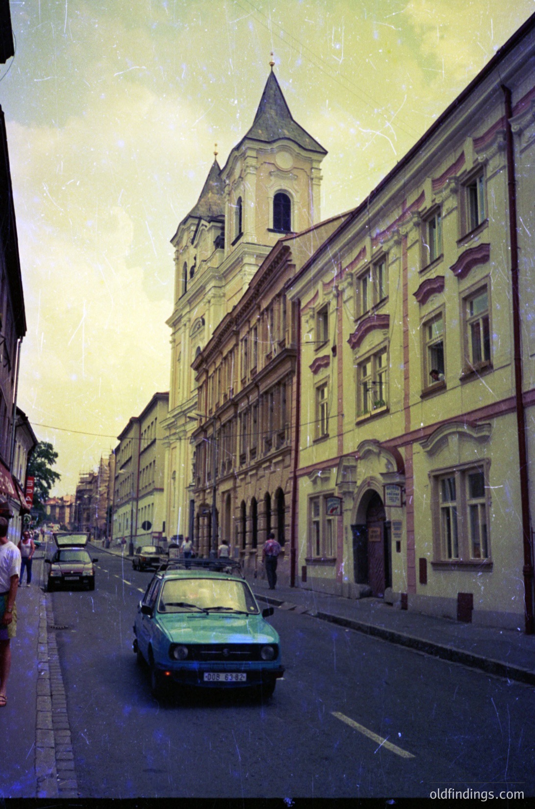 Vintage street scene featuring a row of 19th-century European buildings with classical facades, including a prominent church spire. A green sedan and parked cars line the narrow, slightly inclined road. The architecture includes arched doorways, decorative cornices, and large rectangular windows. The image has a sepia-toned vintage filter. #