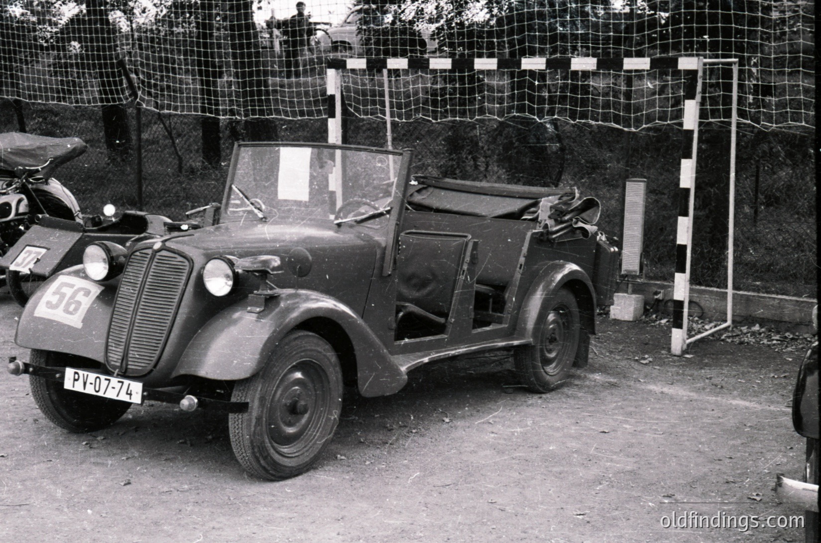 Vintage open-top convertible car, likely a 1930s–1940s European model, parked on gravel with visible license plate "PY-07-74." Sidecar-style rear cargo area and rounded fenders. Barrier net and goalpost frame suggest a sports or rally event setting. Monochrome, high-contrast black-and-white.