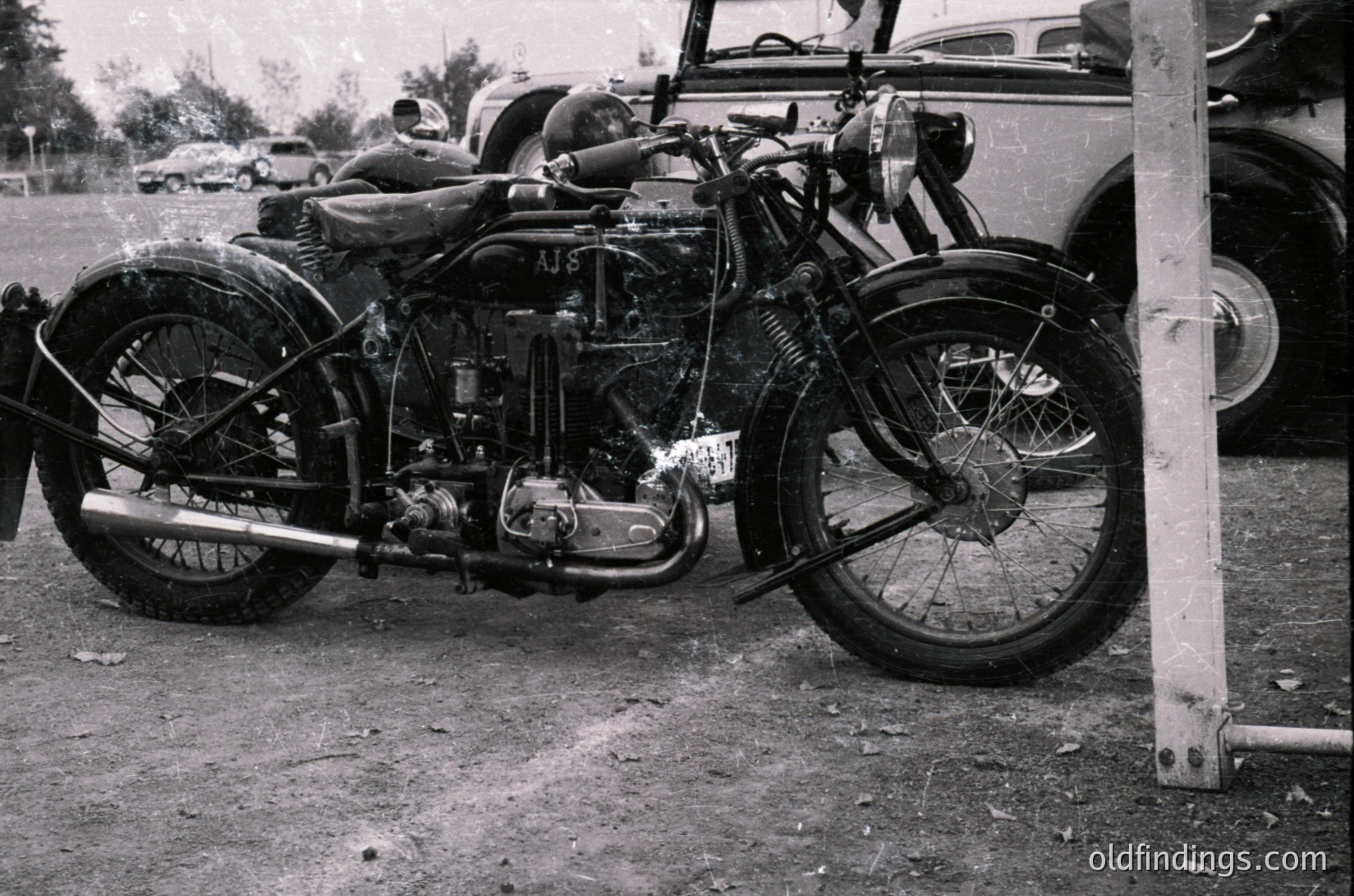Vintage motorcycle with sidecar frame, likely a pre-1940s AJ model, parked outdoors near classic cars. Chrome engine and dual headlamps visible. Rust and wear indicate historical preservation value.
