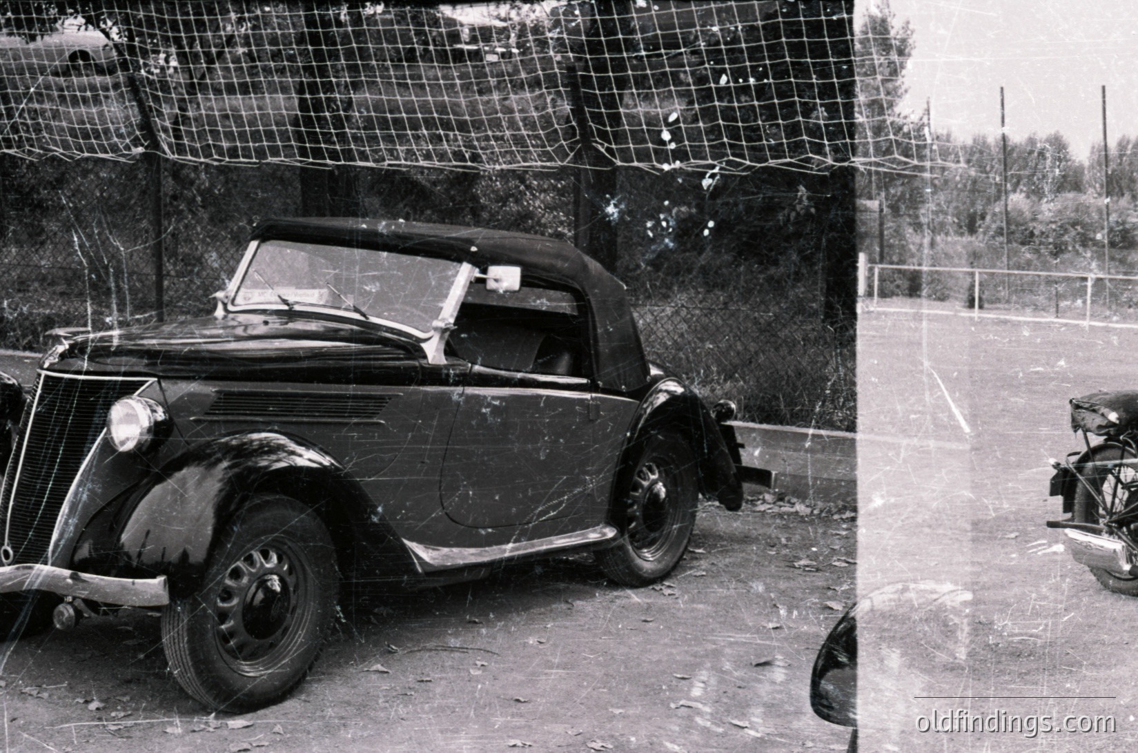 Classic convertible sedan parked beside a tennis court, likely mid-20th century. Distinctive rounded fenders and vintage headlamps suggest a pre-1960s European design. Rust and wear indicate long-term exposure. Tennis net and court markings visible in background.