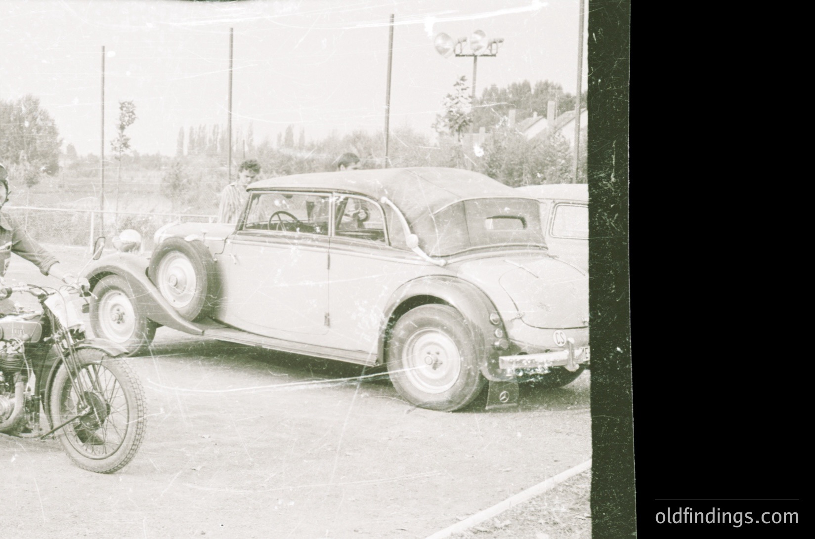 Vintage black-and-white photo of a classic 1930s–1940s roadside scene: a closed-couple car (likely a Ford or Chevrolet) parked beside a motorcycle with sidecar. Rural setting with utility poles, trees, and a distant fence.