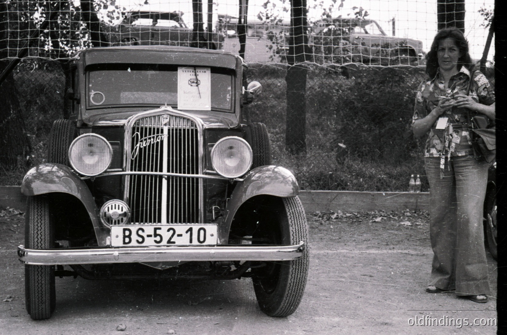 Classic 1930s-era vintage car (BS-52-10) parked beside a chain-link fence, likely Eastern Bloc-era license plate. Woman in patterned blouse and wide-legged pants stands nearby, holding a lanyard. Rustic, outdoor setting with industrial backdrop.