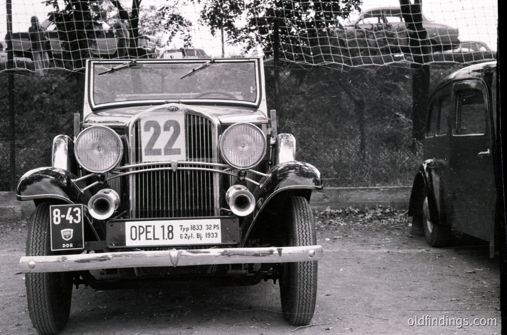 1933 Opel 1.8L Typ 1033 race car (-43) in vintage rally setup. Rounded headlights, wire-spoke wheels, and racing number 22 prominently displayed. Fenced background suggests event or track.