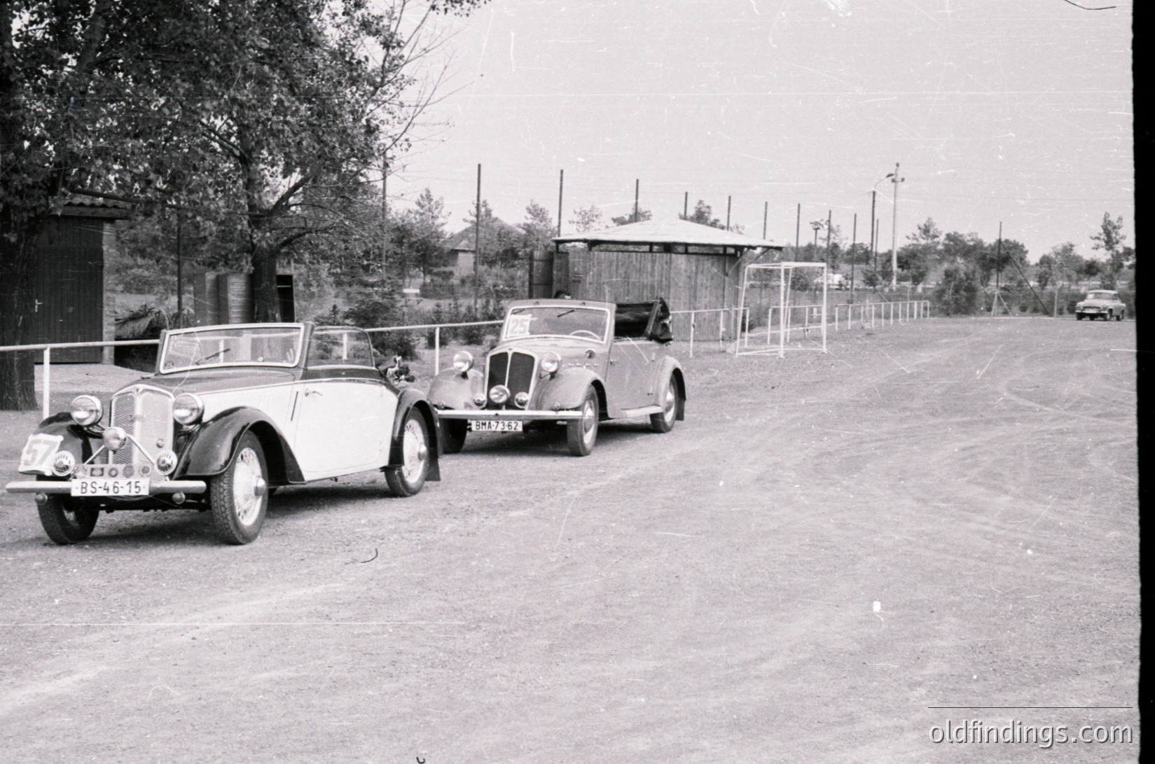 Vintage roadside scene featuring three classic convertibles parked in a row, likely mid-20th century (1940s–1950s). Distinctive rounded fenders and chrome detailing on lead car suggest a pre-war European design. Guardrail and small kiosk in background indicate a toll or checkpoint area.