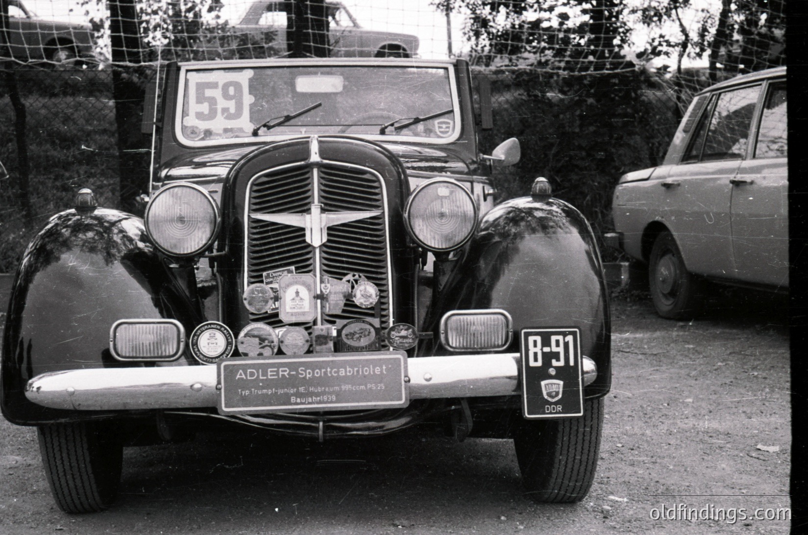 Classic 1930s–1940s Adler Sportcabriolet (Type Trumpf Junior) with DDR license plate "8-91" and race number "59" on hood. Chrome grille, dual round headlights, and vintage racing badges visible. Rustic outdoor setting with modern cars in background.