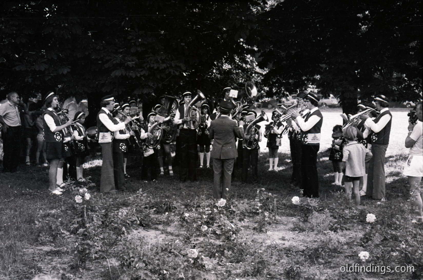 A mid-20th century funeral procession with a brass band playing at gravesites. Uniformed musicians in 1950s-60s attire, including hats and sashes, flank a central figure in formal wear. Graves adorned with white flowers and wreaths. Crowd of mourners, including children, observes solemnly under shaded trees.