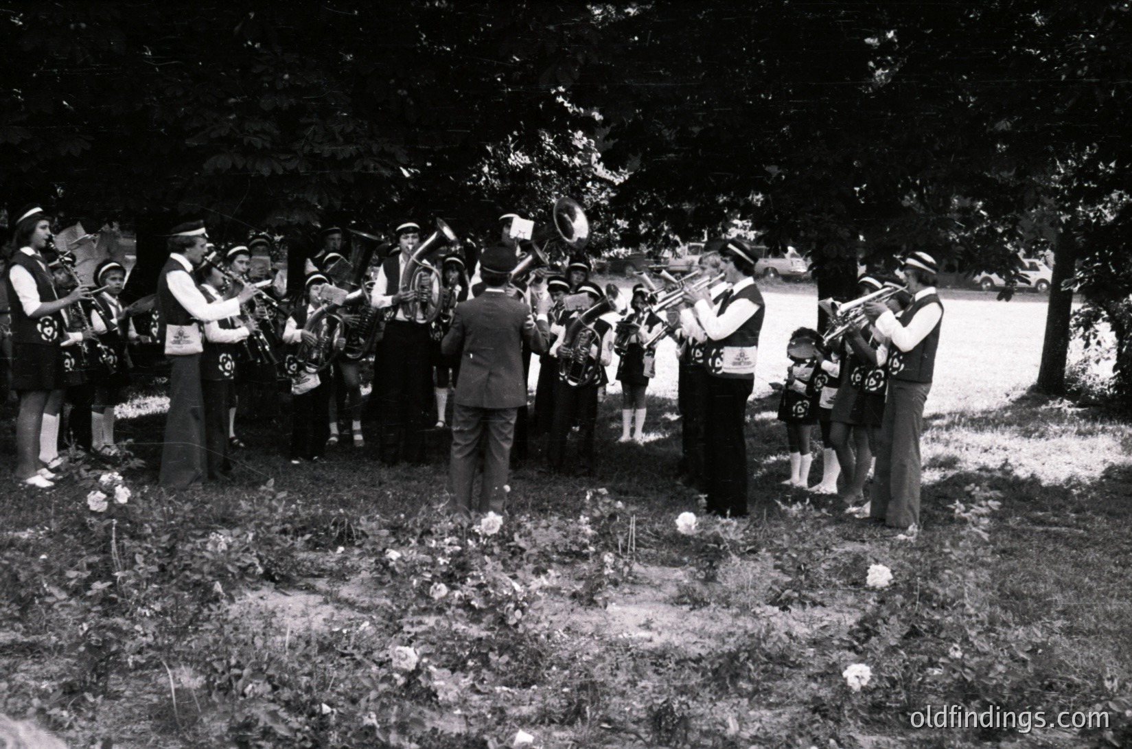 A black-and-white photograph captures a solemn outdoor funeral procession, likely from the **1950s–1960s**. A brass band in formal attire plays at a gravesite, surrounded by mourners—including a child in a dark dress and an adult in a suit. Floral tributes and a simple cross mark the grave. The setting appears rural or suburban, with trees framing the scene.