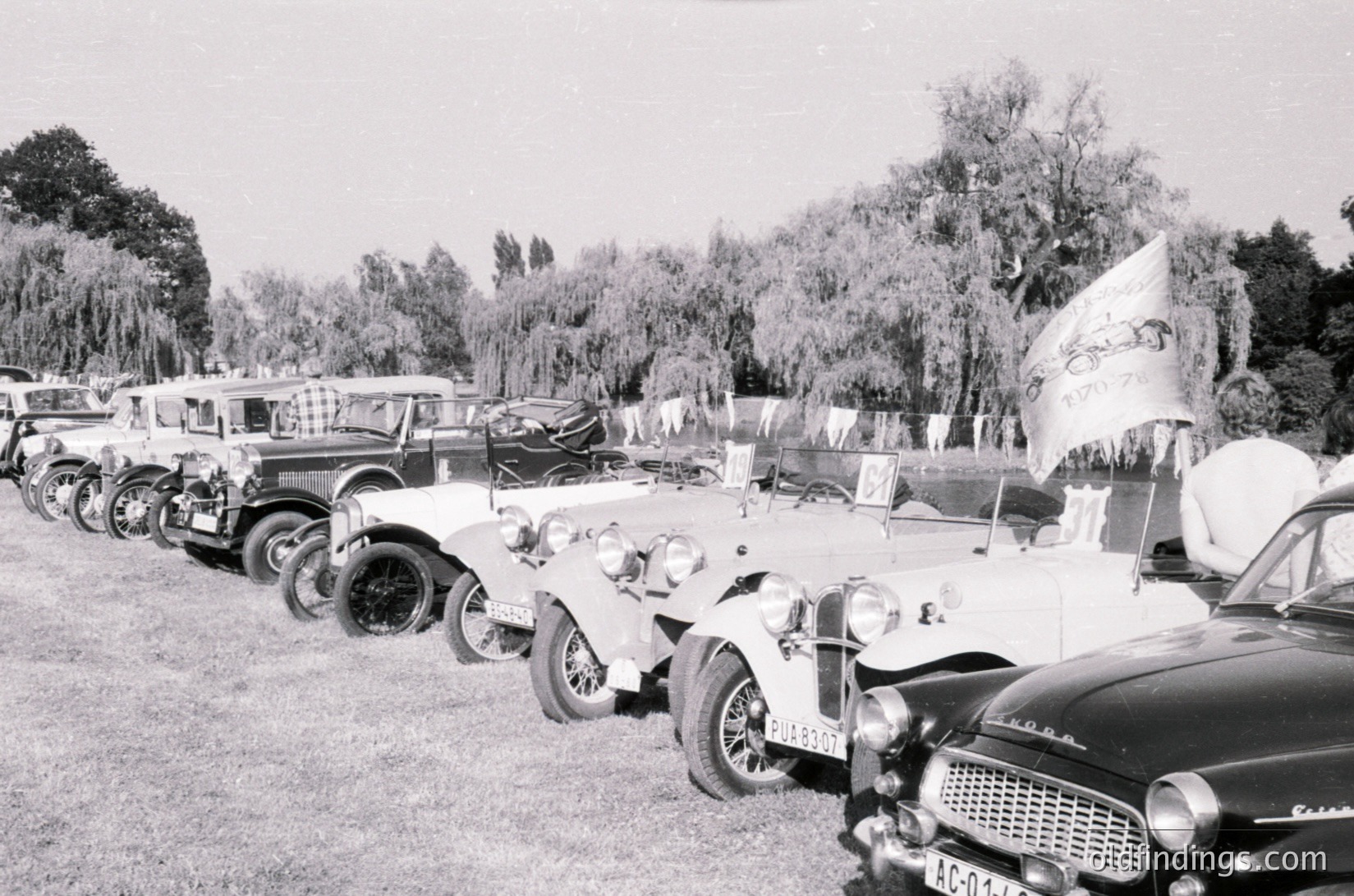 Classic vintage cars lined up in an outdoor setting, likely a 1960s automotive show or rally. Visible models include a vintage Škoda with "AC-03" plate and a pre-war Mercedes-Benz 300SL. Lush trees and a banner in the background suggest a park or event venue. #ŠkodaHistory