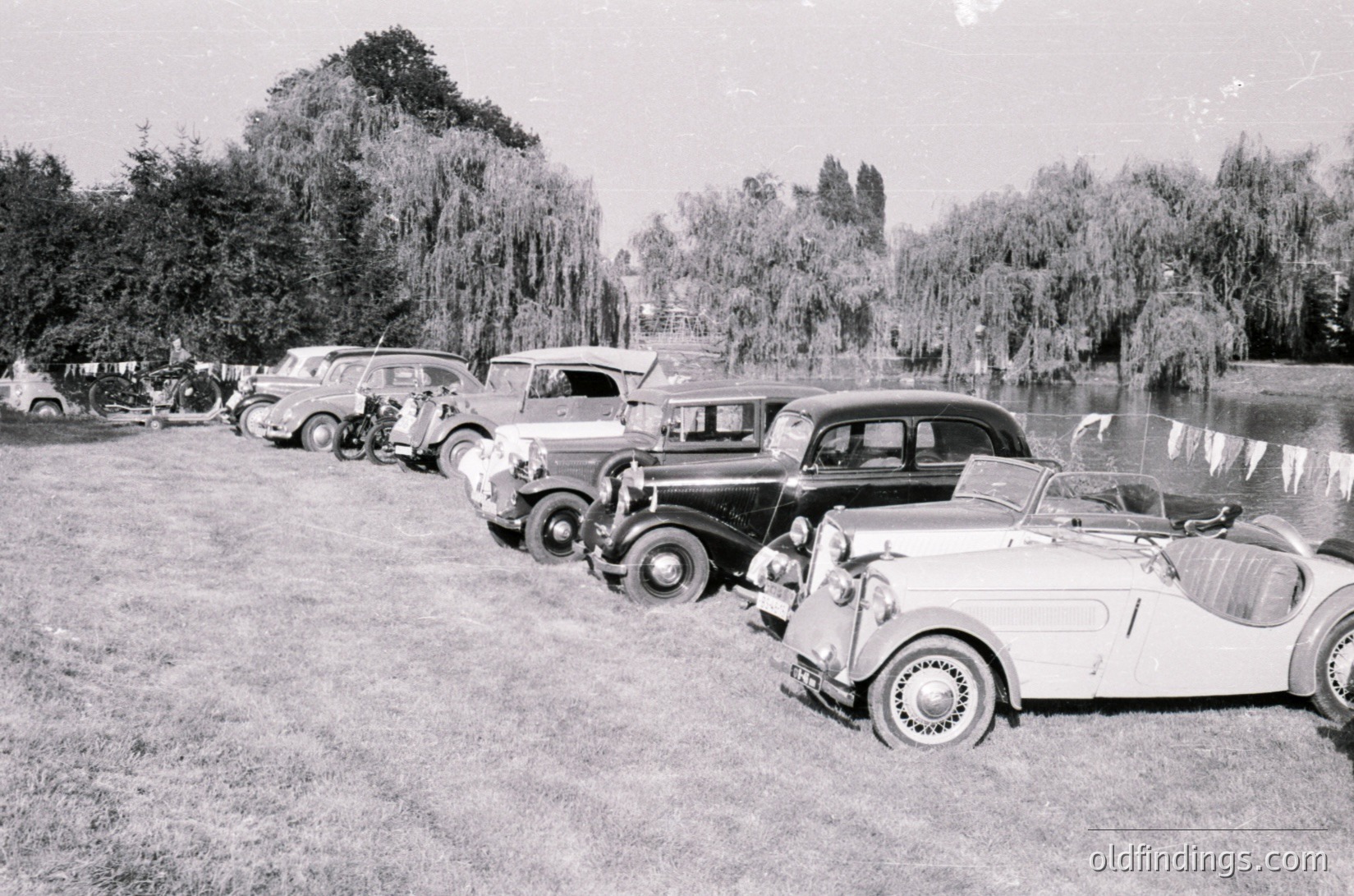 Vintage car lineup by a serene lakeside, featuring classic 1930s–1940s models including a convertible with whitewall tires and a streamlined sedan. Lush weeping willows frame the scene, suggesting a park or lakeside gathering. Black-and-white monochrome enhances nostalgic atmosphere.