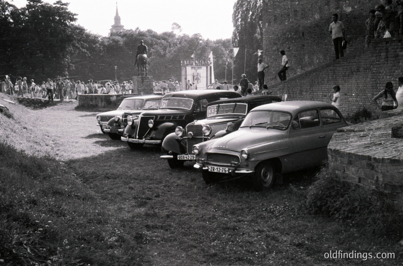 Classic 1960s European rally scene: vintage cars (Volvo PV544 & Fiat 1200) parked on gravel beside a stone-walled hillside. Crowd gathers on steps near a church spire, watching event.