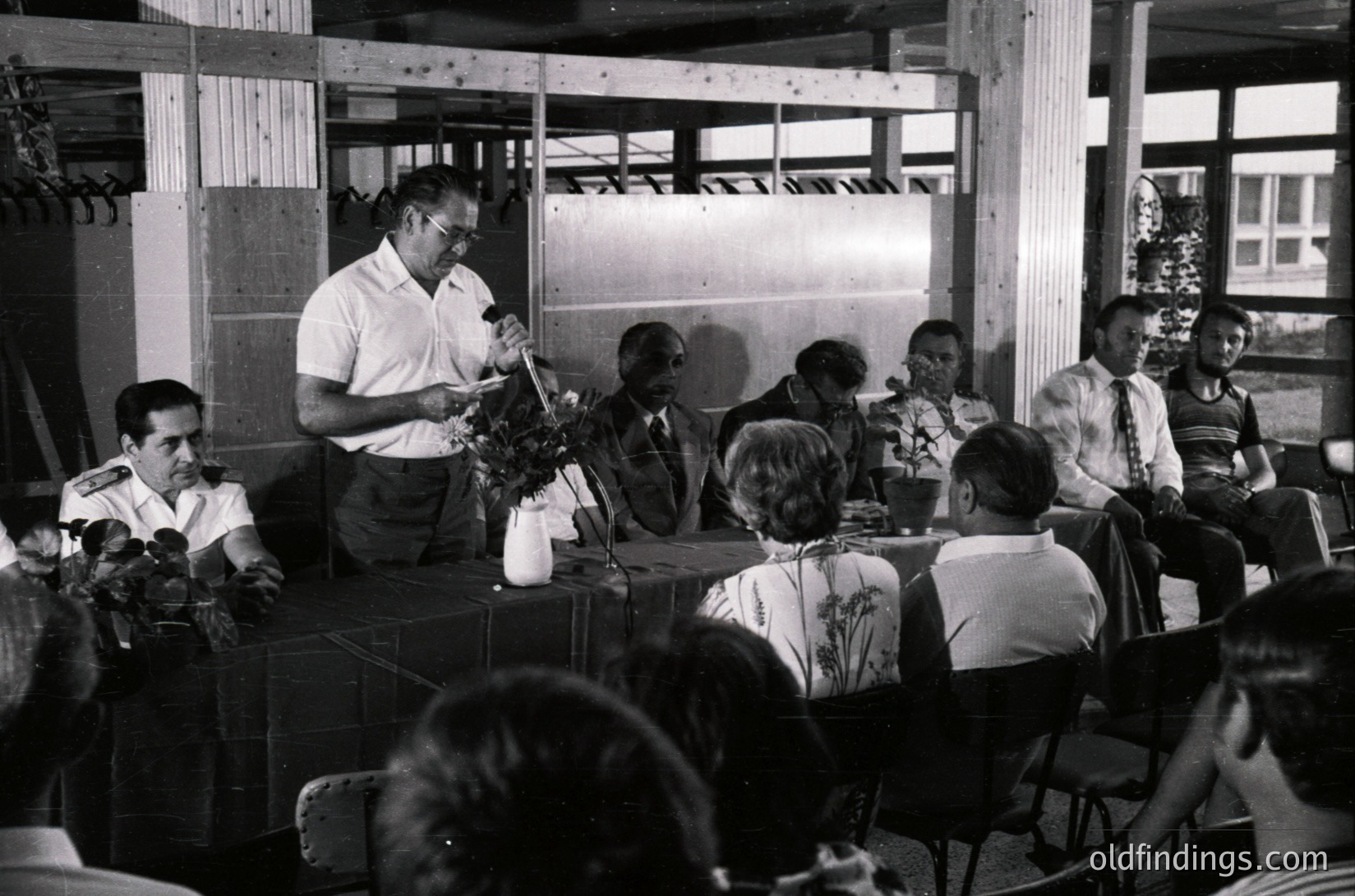 Indoor formal gathering featuring a seated panel discussion or ceremony. A man in a white shirt and tie stands at a table holding a bouquet, likely presenting an award or honor. Six seated individuals in formal attire—ties, blazers, and dresses—face the speaker. Wooden paneling and simple wooden chairs suggest a mid-century institutional or cultural venue. Crowd in background indicates public attendance.