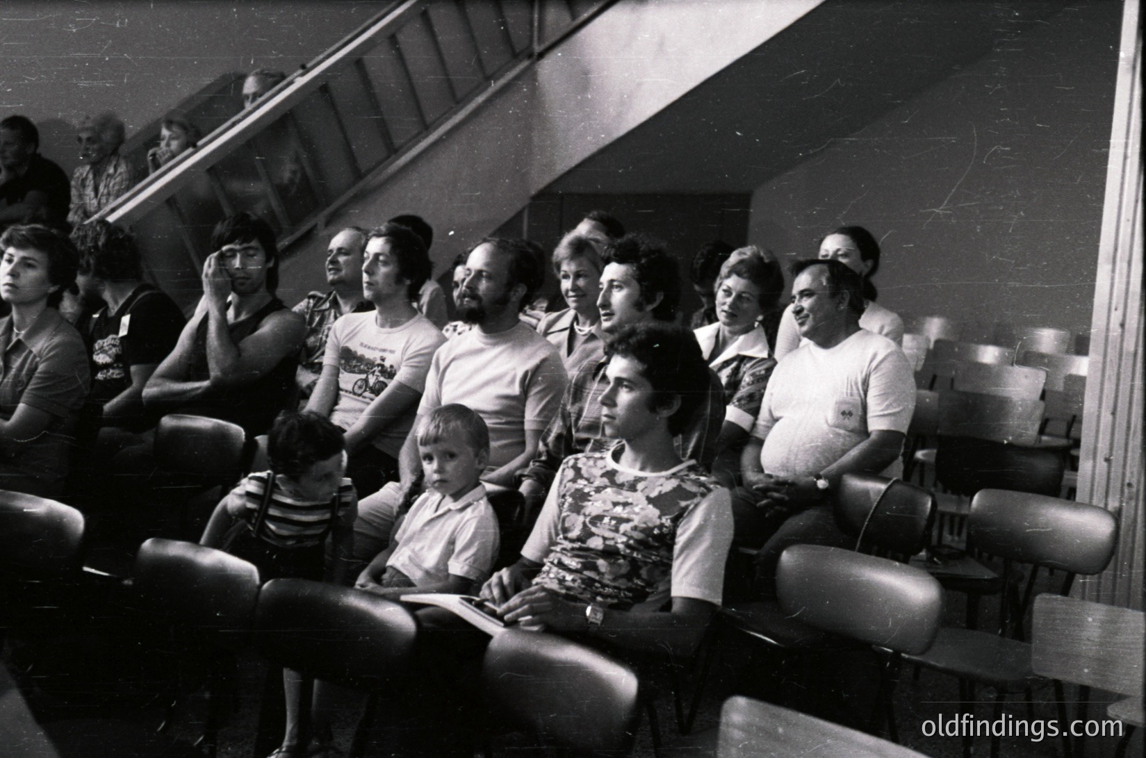 A black-and-white photo of a mixed-gender audience seated in tiered theater-style seating, likely from the 1970s–1980s. Casual attire includes patterned blouses, button-down shirts, and a child in a striped shirt. The setting appears to be a public indoor venue, possibly a cultural or community event. The lighting is dim, emphasizing the focus on the audience.