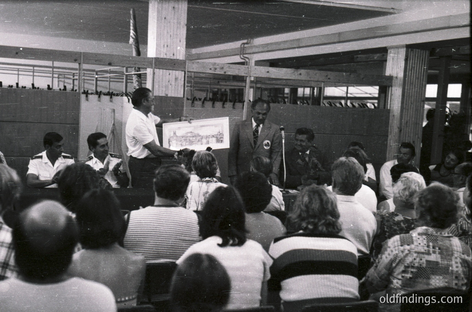 Indoor lecture hall with audience seated in rows facing two speakers. One speaker holds a large poster of a coastal landscape, likely for educational or touristic purposes. Attendees wear 1960s–1970s casual and semi-formal attire. Industrial-style lighting and exposed beams suggest a functional, utilitarian space.