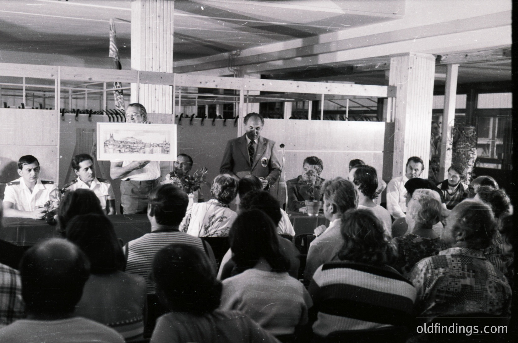 Black-and-white photo of a mid-20th-century lecture or meeting in an industrial hall. A speaker at a podium holds a poster with a building sketch, addressing a seated audience of ~30 people. Wooden beams and exposed trusses frame the scene, suggesting a utilitarian or temporary venue. Attendees wear 1960s-70s clothing (striped shirts, blazers).