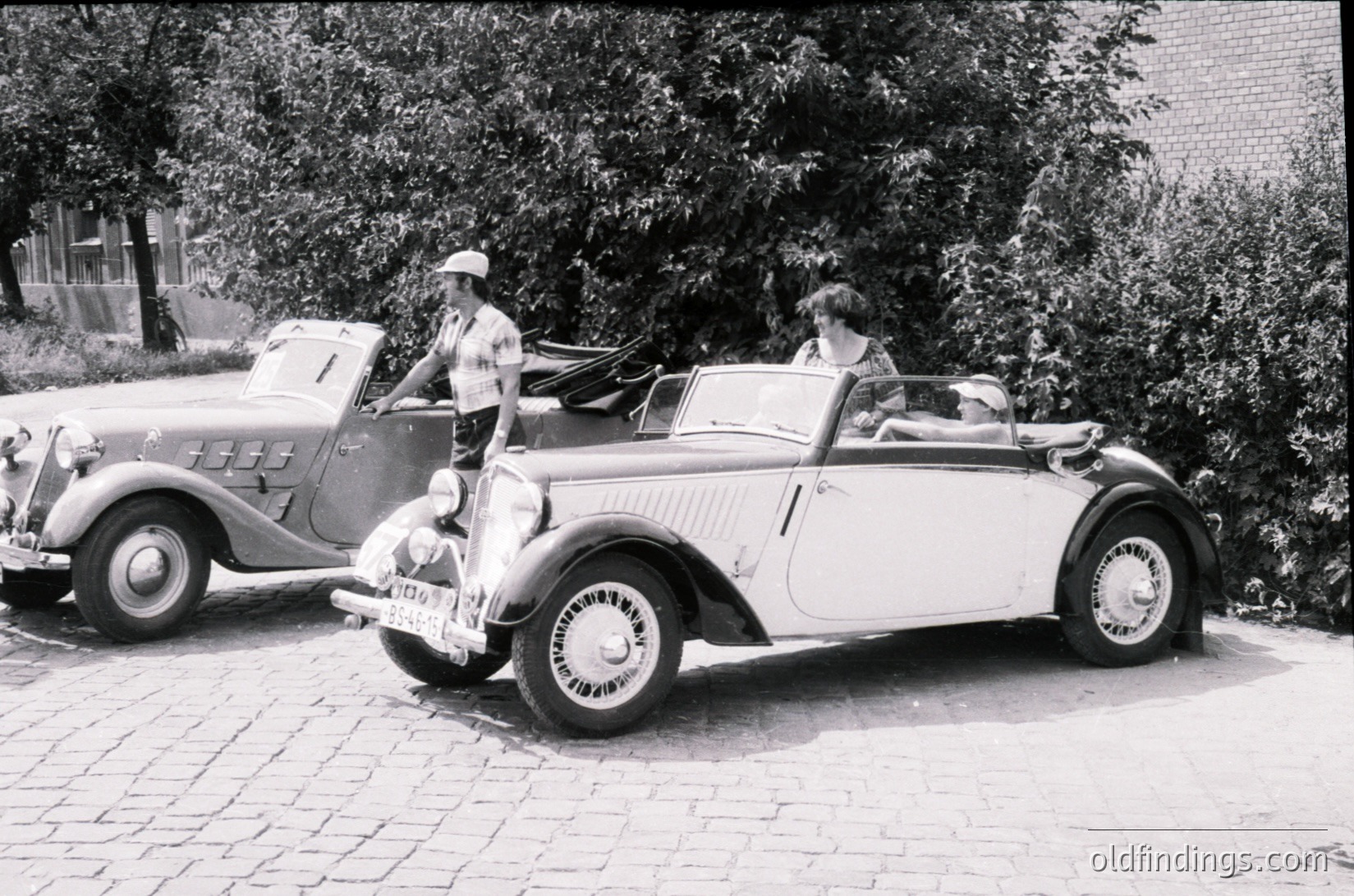 Vintage 1930s–1940s roadside scene featuring a white convertible with streamlined bodywork and wire-spoke wheels, parked beside a classic sedan. A man in a hat and light shirt stands beside the convertible, while a woman in a dress sits inside. Lush greenery and cobblestone pavement enhance the nostalgic atmosphere.