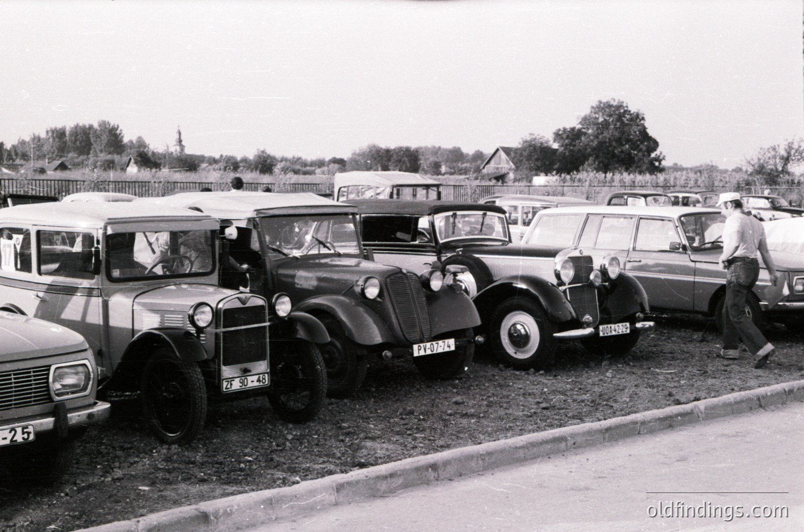 Vintage lineup of classic cars in a rural setting, likely Eastern Europe, 1960s–1970s. Notable vehicles: Soviet-made GAZ-51 truck, pre-war European sedans, and a 1950s-era station wagon. Men in casual attire inspect or stand near vehicles. Fenced residential area and trees in background.