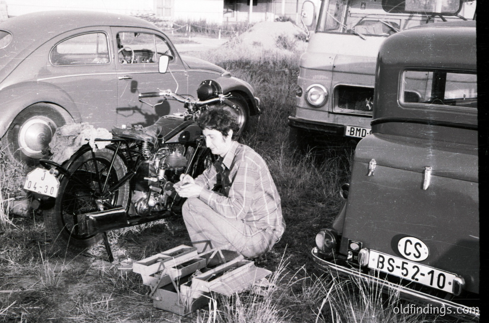 Mid-century mechanic tinkers with a vintage motorcycle engine in an open field beside classic cars (BS-52-10, BMW license plates). Overalls and toolbox suggest 1950s–1960s European repair culture.