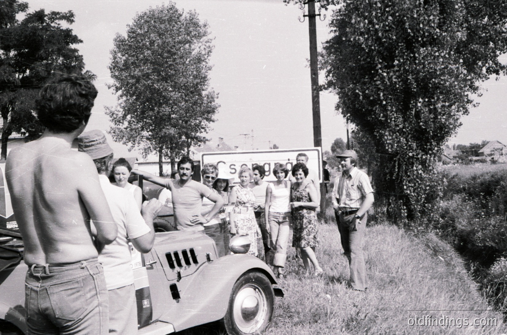 Vintage black-and-white photo of a group posing with a vintage truck, likely 1960s–1970s. Casual summer attire (shorts, hats) suggests rural or roadside gathering. Sign in background reads "SOS" with blurred faces. Open countryside with trees and utility poles.