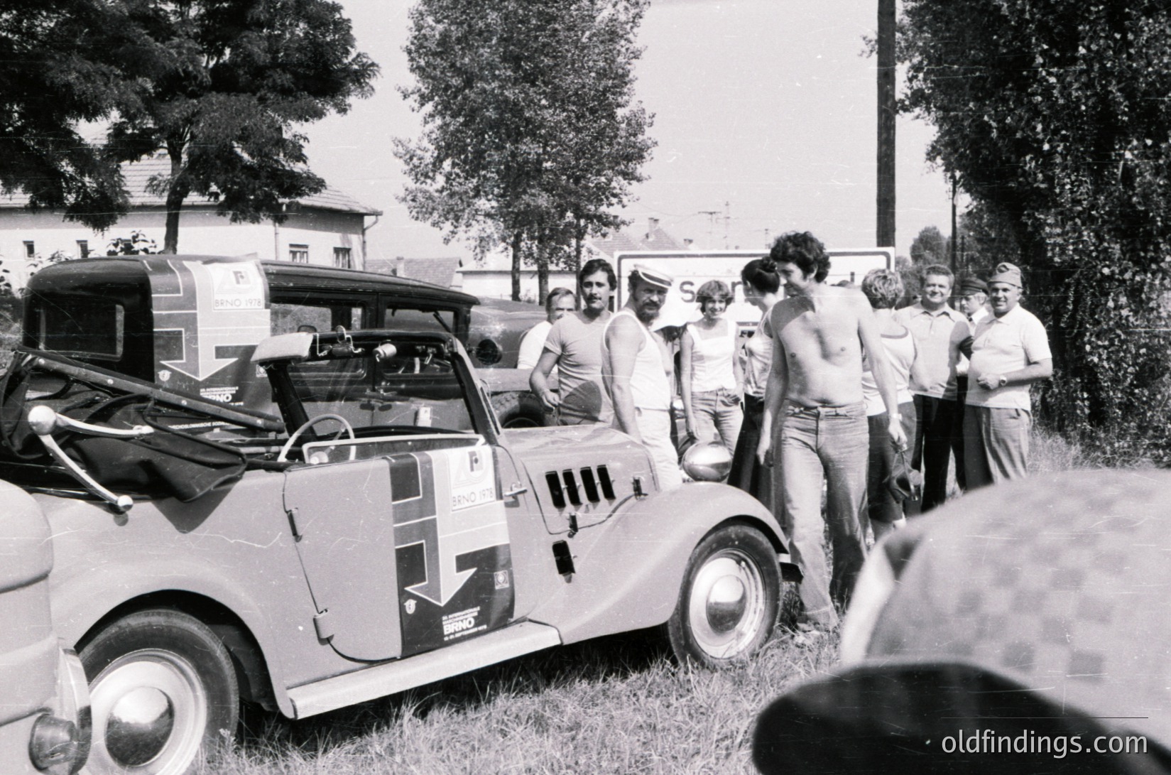 Vintage rally car with sponsor decals and mud splatter, surrounded by a group of casually dressed spectators in 1970s outdoor setting. Classic rally scene with trees and grassy area.