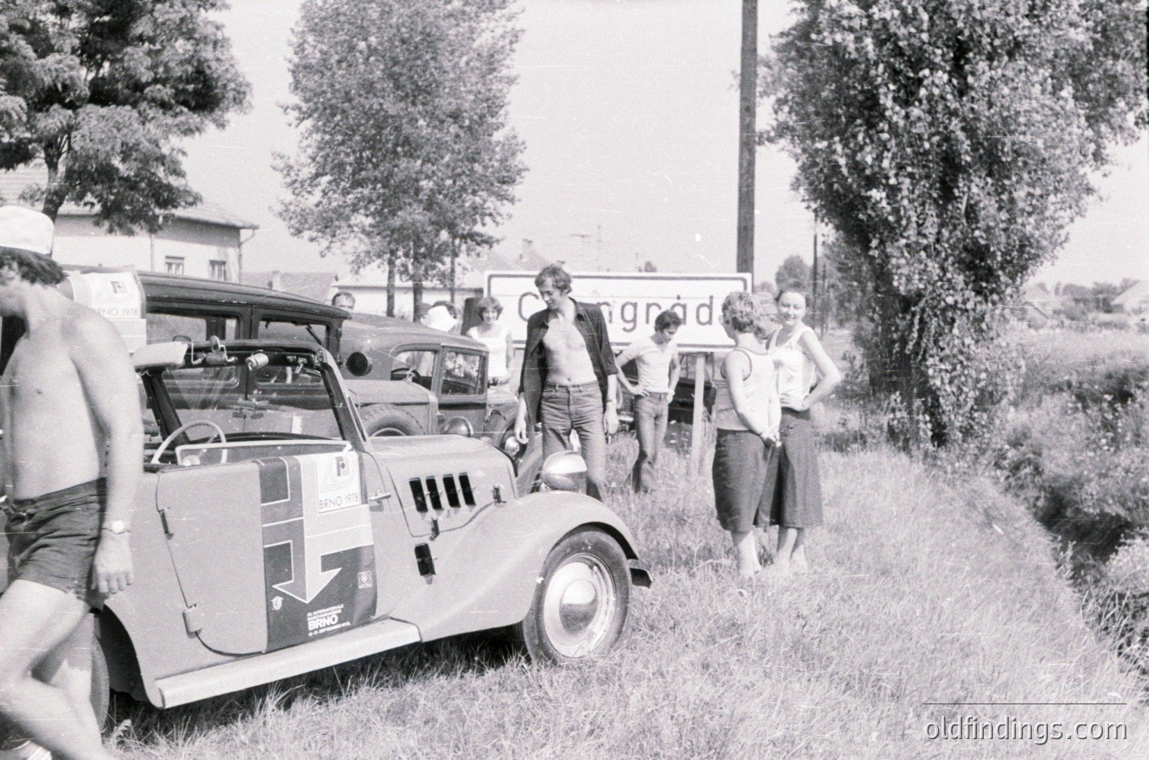 Vintage rally car with sponsor decals paused beside a rural roadside, surrounded by onlookers in 1960s-era clothing. Signage reads "Congratulations" in blurred text. Lush greenery and trees frame the scene, suggesting a countryside setting.