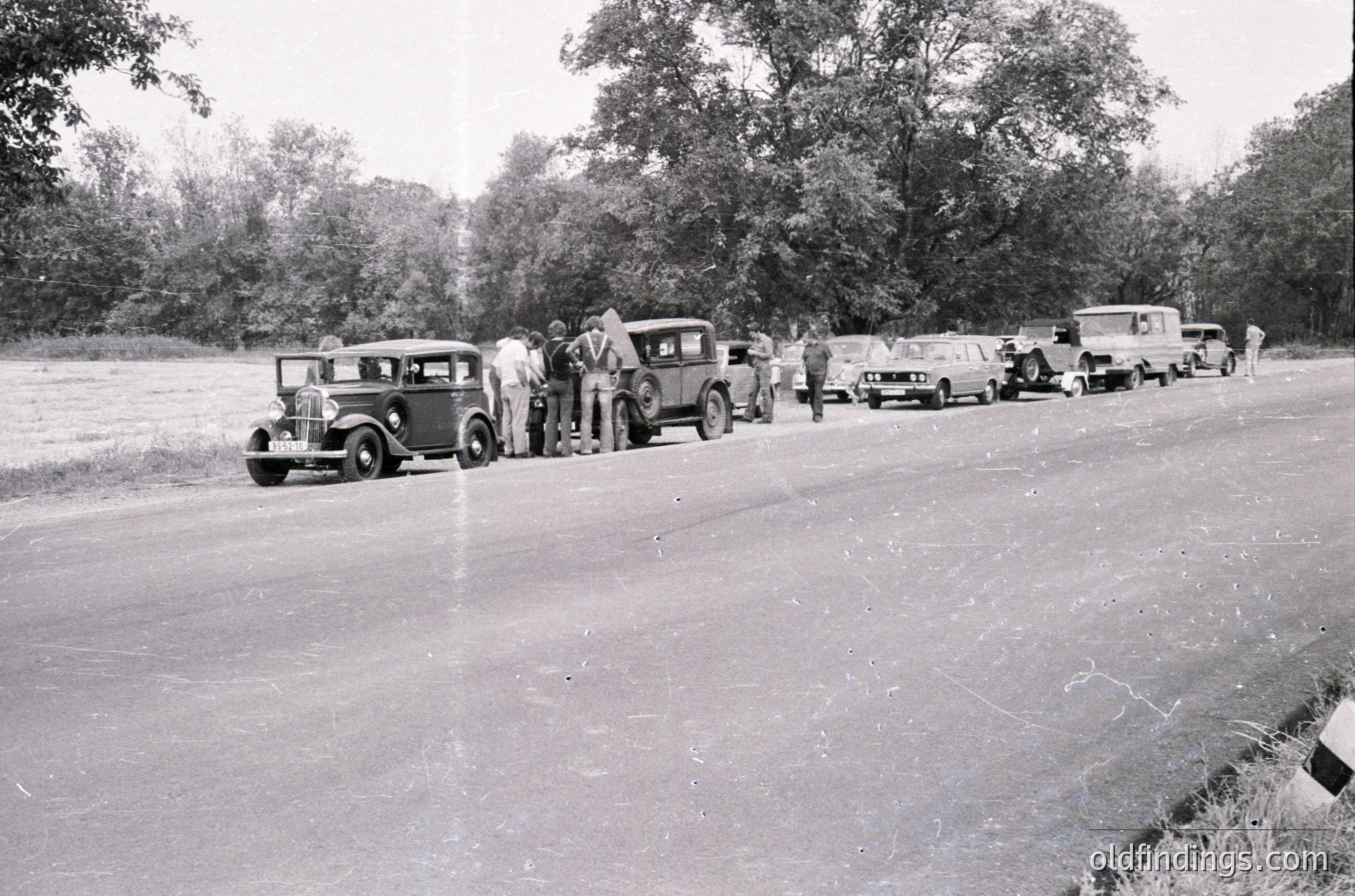 Vintage roadside gathering of vintage cars (1930s–1940s) lined up on a rural road, surrounded by dense trees. Group of people in period attire inspecting vehicles. Black-and-white photo captures mid-century automotive culture and social interaction.