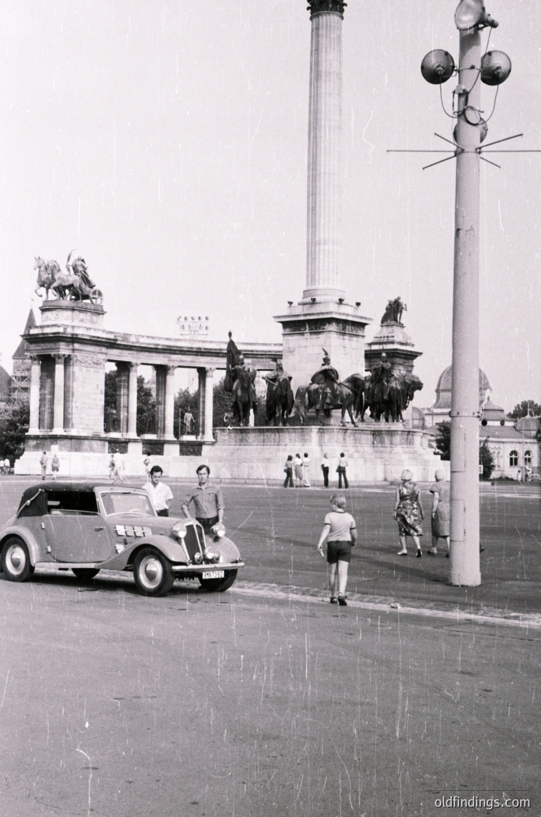 1950s-era monument with equestrian statues and classical columns in Budapest, Hungary. A vintage car with passengers poses near Heroes' Square, flanked by pedestrians in mid-century attire. Architectural grandeur meets urban daily life.