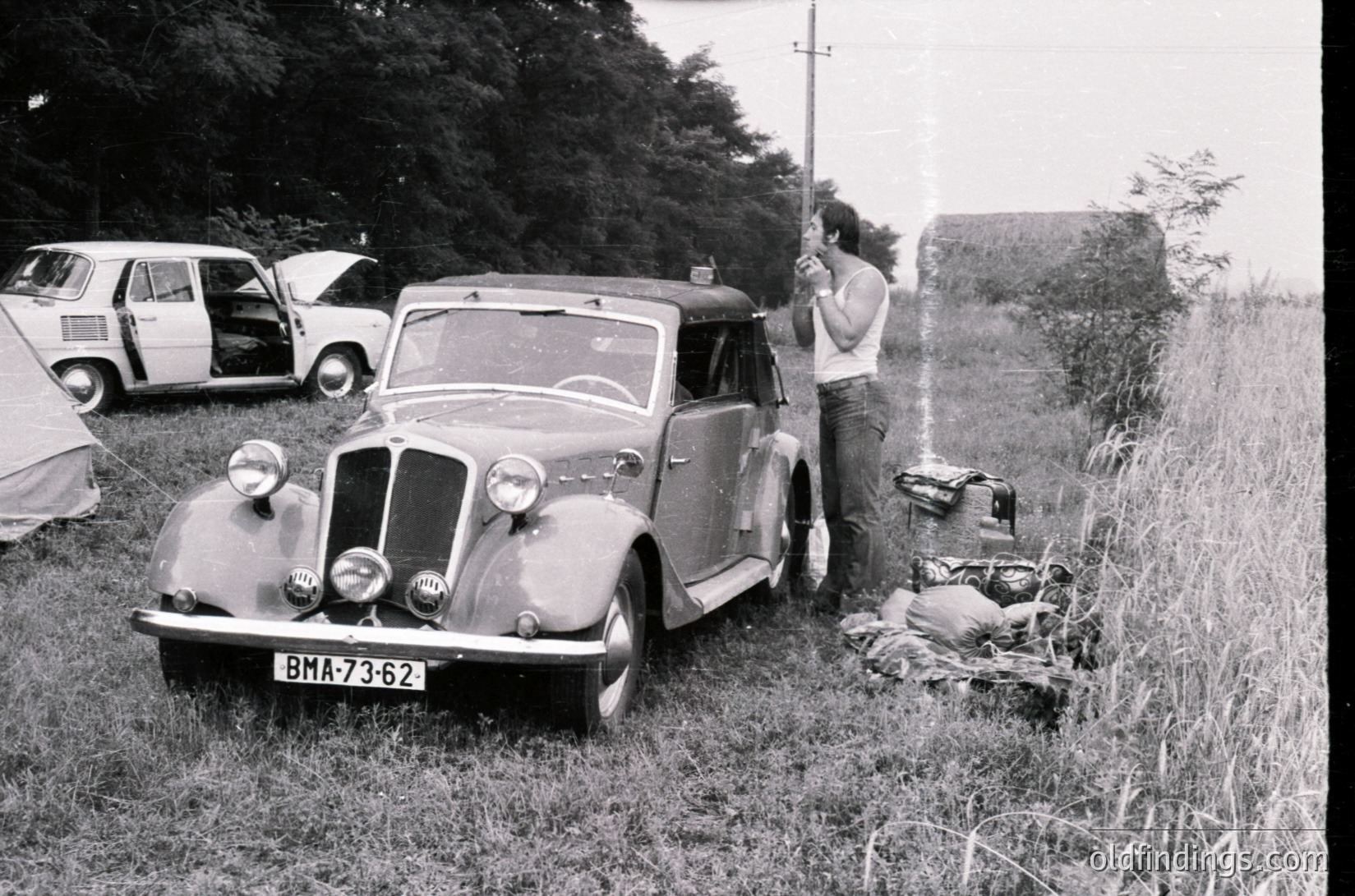 Vintage 1950s-era sedan (BMA-73-62) parked in grassy field with classic rounded headlights and chrome accents. Man in white shirt adjusts camera near overgrown concrete structure. Other vintage vehicles in background. Rural or outdoor event setting.