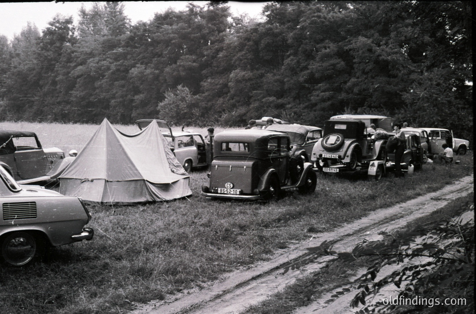 Vintage camping scene with classic cars and tents along a lakeside dirt road, mid-20th century. Vehicles include a vintage truck, sedan, and station wagon parked near a canvas tent. Dense forest and water body in background.