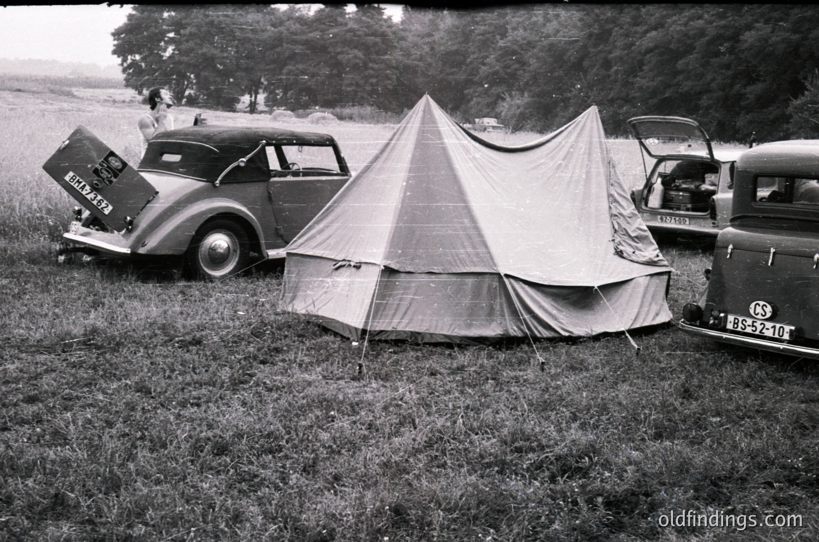 Vintage 1950s-era camping scene with classic cars and canvas tent in open grassy field. Pre-war sedan (1940s) with visible license plate "BUC 277" parked beside a large, peaked canvas tent. Another car (1950s) with "CS 52-10" plate in background. Rural, mid-century outdoor recreation.